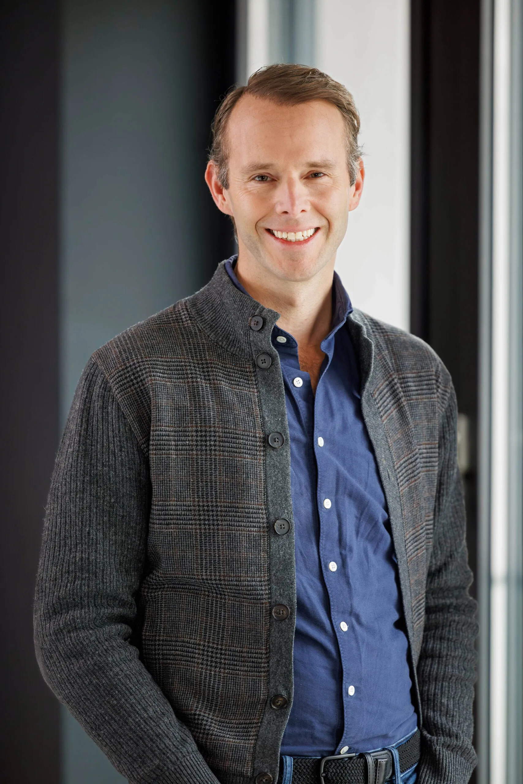 Portraits von der Geschäftsführung, ein Mann mittleren Alters mit braunem kurzem Haar. Er trägt ein blaues Jeanshemd, eine grau karierte Strickjacke und er lacht in die Kamera. Im Hintergrund dunkle, unscharfe Strukturen in Schwarz und Grautönen.