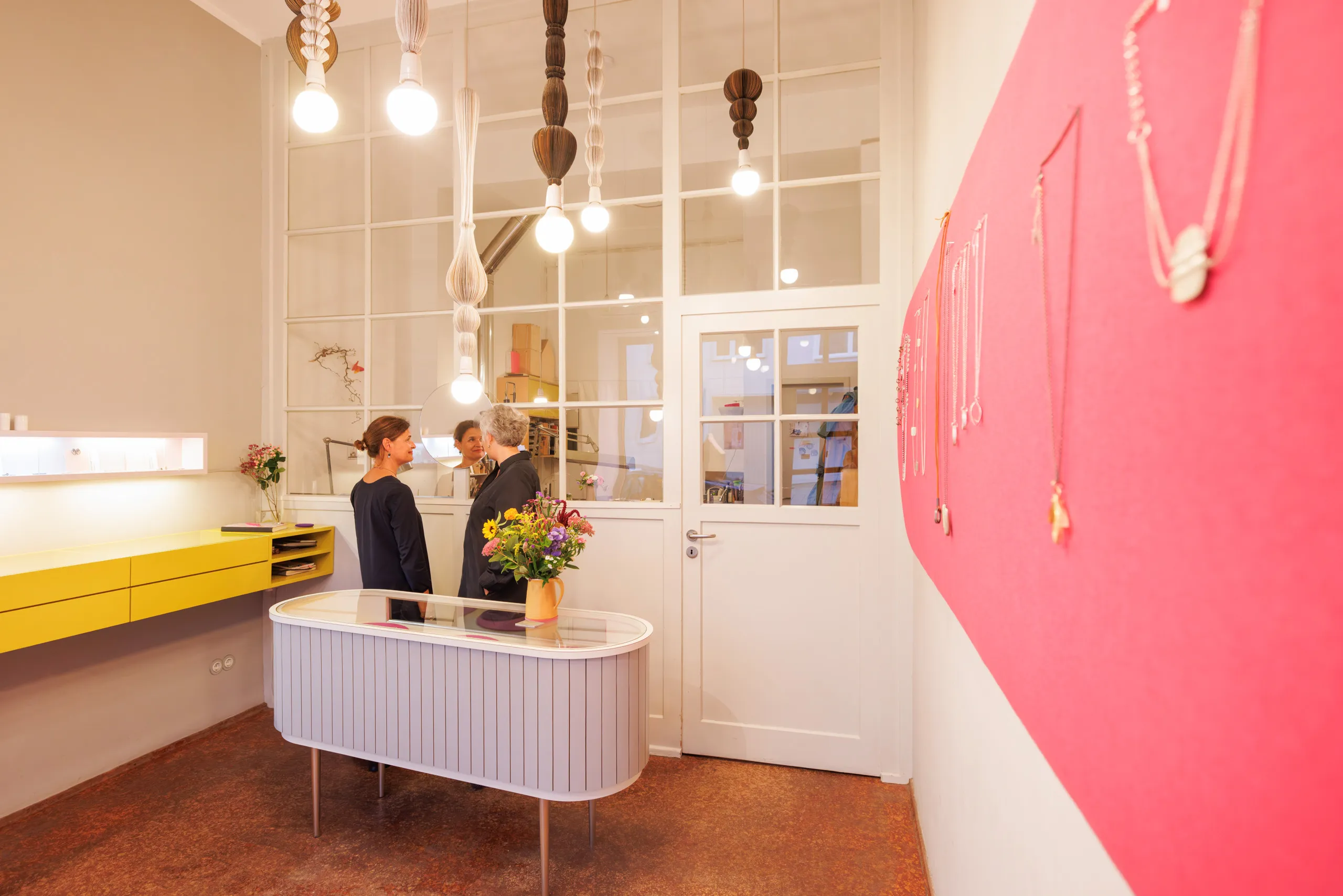 Blick in den Schmuckladen, rechts eine rosa Wand mit Schmuck und eine weiße Schmuckvitrine mit einer Blumenvase zu sehen. Da hinter stehen zwei Goldschmiedinnen in dunklen Kleidern und unterhalten sich, viele, moderne Lampen an der Decke und links befindet eine gelbe Vitrine an der Wand. Der Fussboden hat eine rotbraune Färbung.