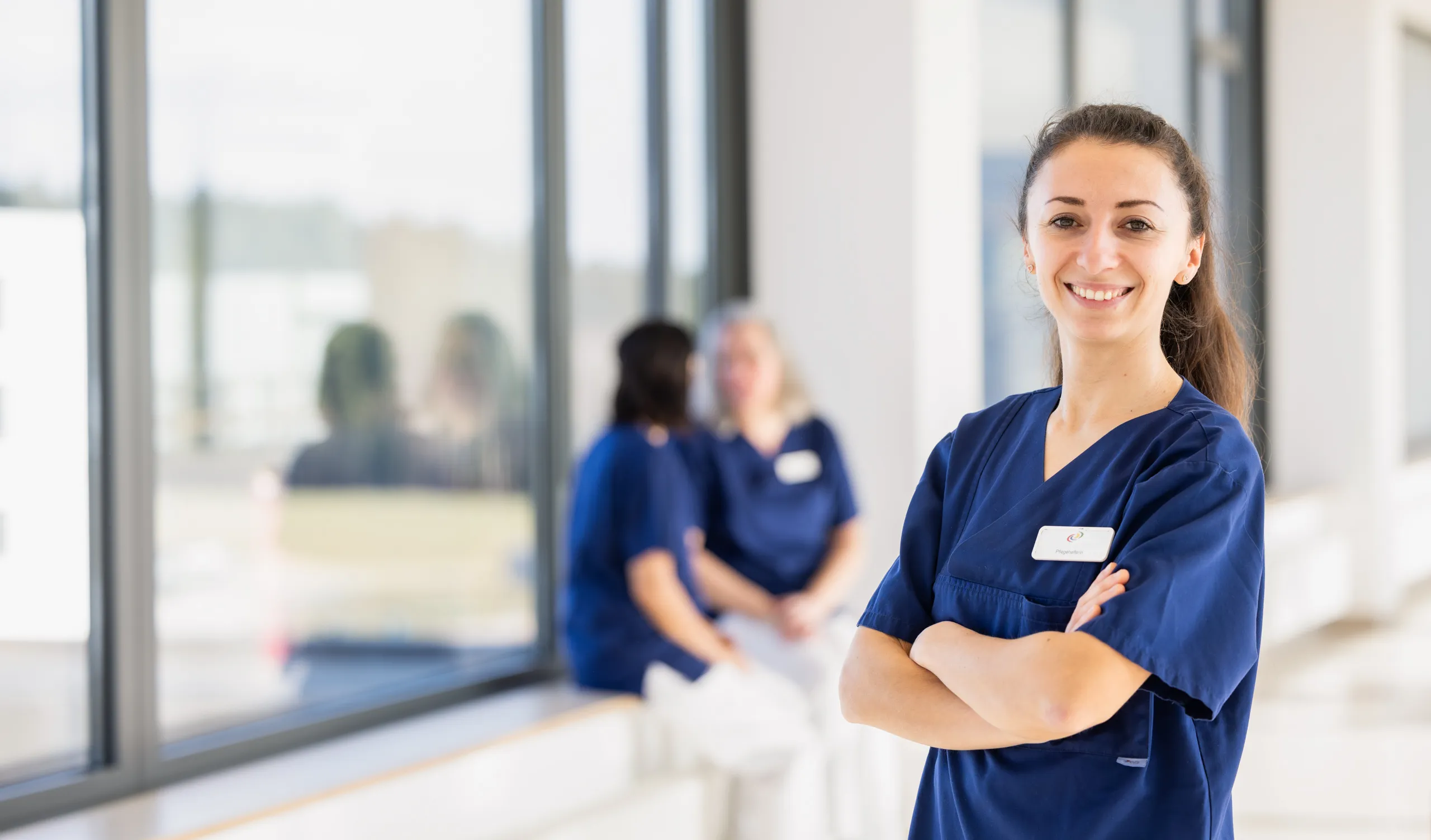 Das Foto zeigt einen weißen Kliniken, mit grauen Fensterrahmen und ist lichtdurchflutet. Ganz rechts im Bild steht eine Krankenschwester in einem dunkelblauen Kasack, mit verschrenkten Armen und sie lacht in die Kamera. Sie hat braune, lange Haare zu einem Zopf gebunden. Mittig im Foto sitzen zwei Pflegerinnen auf einer Fensterbank in der Unschärfe. Beide Frauen tragen weiße Hosen und blaue Kasack. Sie unterhalten sich. Die Dame links hat dunkle, schulterlange Haare und die andere Dame hellgraue, schulterlange Haare.