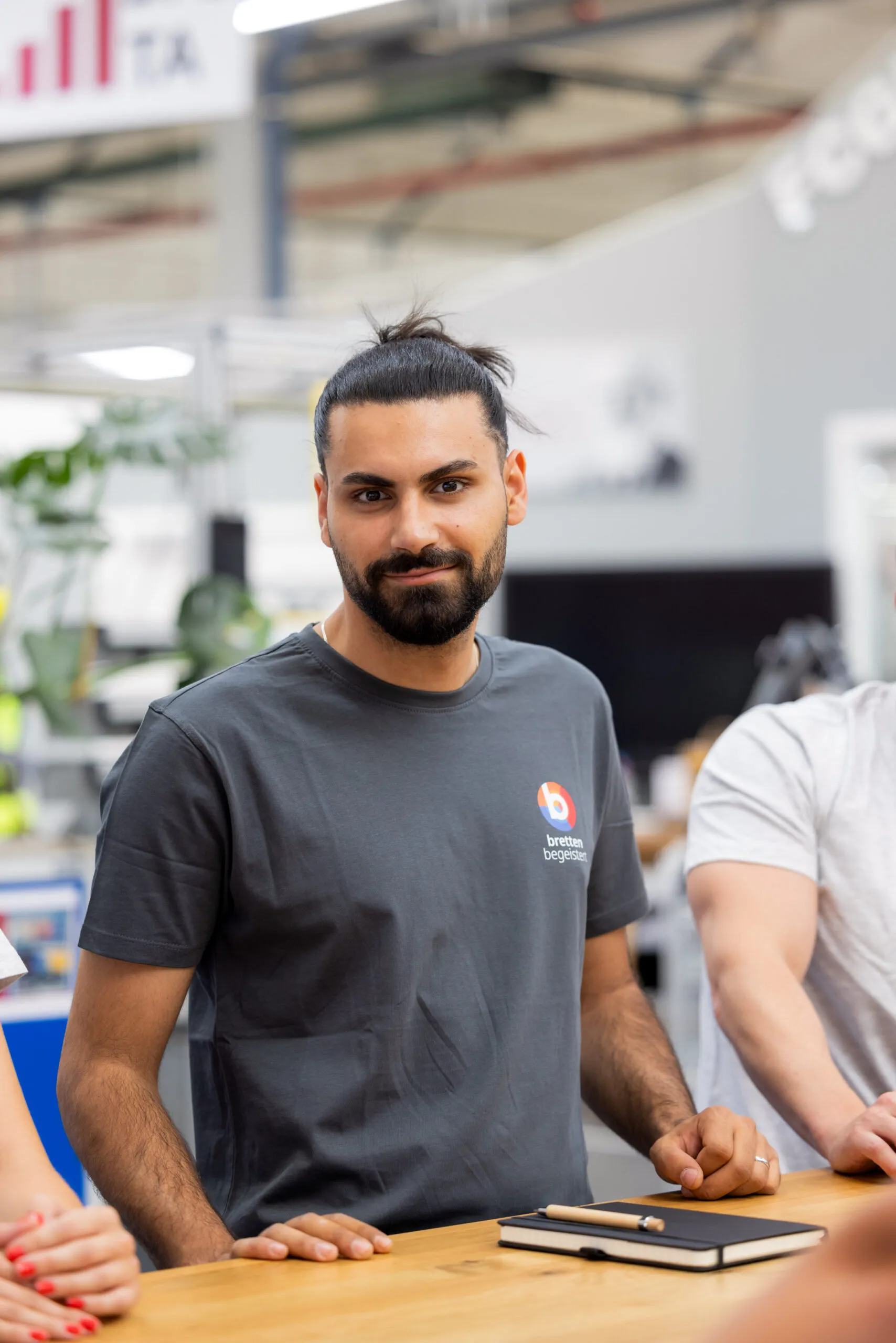 Ein junger Mitarbeiter mit dunkelhäutiger Haut, schwarzem Bart und Haar, zurück zu einem Zopf gebunden, steht mit dem Oberkörper zu sehen an einem Holztisch. Er trägt ein dunkelgraues „Bretten begeistert“ T-Shirt. Links und rechts von ihm im Anschnitte andere Personen. Im Hintergrund unscharf eine Halle mit Pflanzen und einem Bildschirm zu sehen.