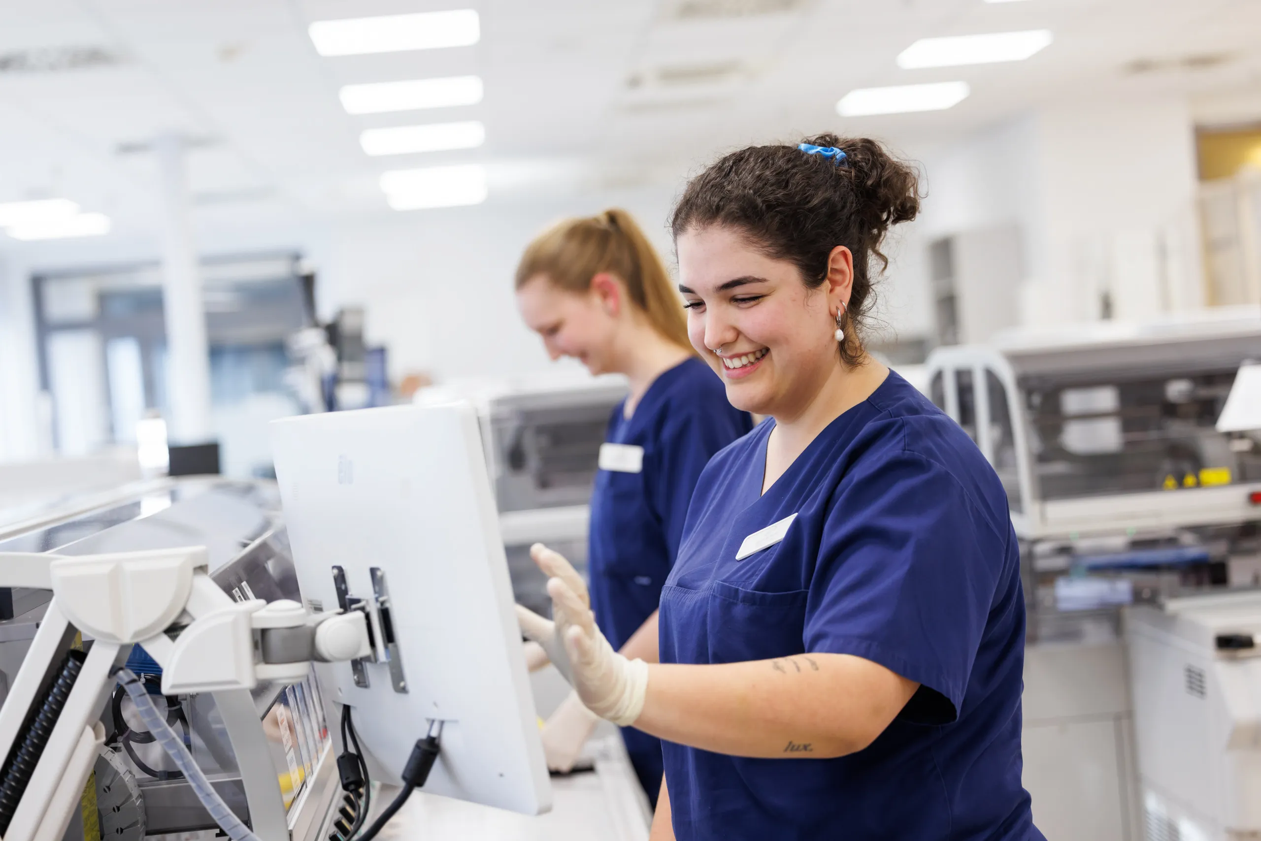 Zwei Mitarbeiterinnen des Klinikums stehen an einer Maschine in einem Labor. Sie tragen beide dunkelblaue Kasacks, arbeiten, schauen nicht in die Kamera und lachen dabei. die vordere Dame hat lockige schwarze Haar zu einem Zopf gebunden, mehrere silberne Ohrringe, einen Nasenring und ein paar Tatoos auf ihrem Unterarm. Die Zweite ist im Hintergrund unscharf zu sehen, mit dunkelblonden, langen Haaren zu einem Zopf gebunden. Die dunkelhaarige Frau trägt weiße Handschuhe und hat ihren linken Arm an einem Monitor. Im Hintergrund ist unscharf ein heller Laborraum zu sehen.