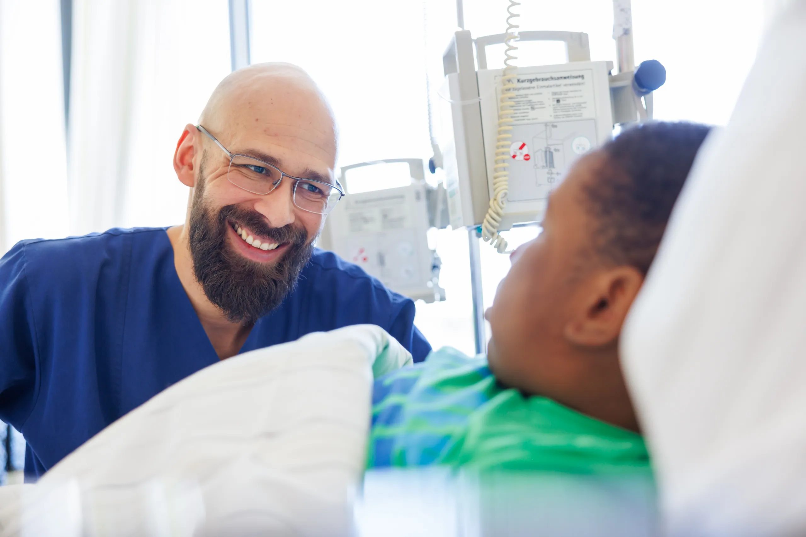 Eine Nahaufnahmen von einem Krankenpfleger und einem farbigen Kind. Der Junge rechts im Bild hat ganz kurze, dunkle Haare, trägt ein grünes T-Shirt und liegt in einem Bett mit weißem Bettzeug. Der Mann links trägt einen blauen Kasack, eine rechteckige Brille, er hat eine Glatze und einen dunkelbraunen Vollbart. Er beugt sich zu dem jungen Patienten und lacht sehr herzlich. Im Hintergrund unscharf ein Infusionsständer mit Kabel und ein überstrahltes Fenster.