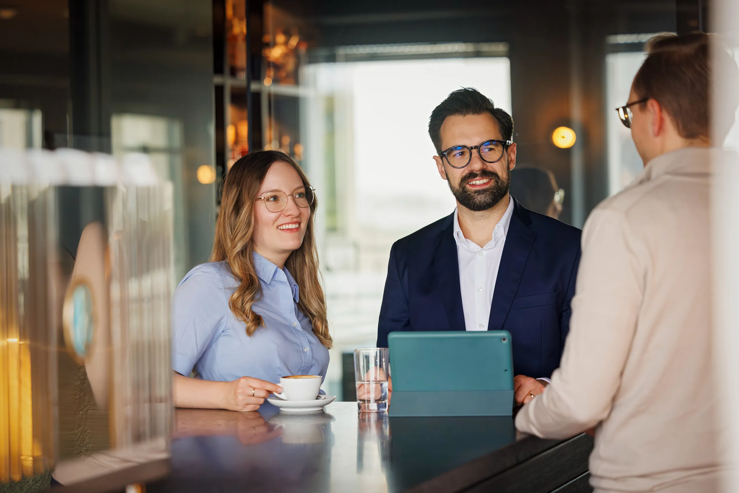 Drei Personen jüngeren Alters stehen an einer modernen, dunklen Cafebar und unterhalten sich. Links steht eine Frau im blauen Hemd, Goldbrille und rotblondem langen Haar mit einer weißen Kaffeetasse. In der Mitte ein Business Mann mit türkisfarbenen Tablet. Er trägt ein dunkelblaues Sakko, weißes Hemd und eine dunkle Brille. Beide schauen zu dem Mann rechts, der unscharf in einem beigen Pullover mit dem Rücken zu der Kamera steht. Er hat kurze braune Haare und eine Brille. Im Hintergrund dunkle Wände mit orangefarbenen Licht und Fenstern.