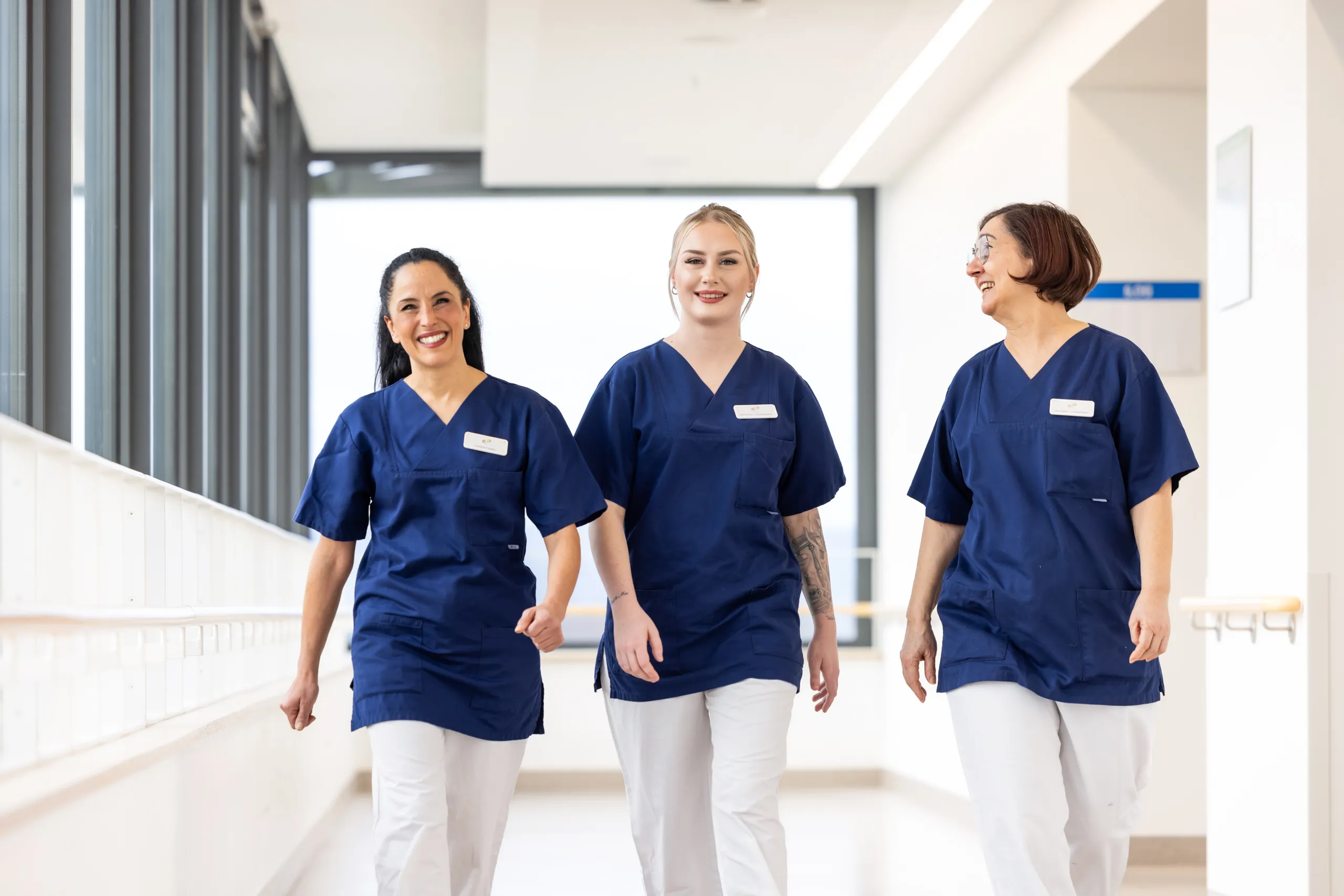 Drei Krankenpflegerinnen gehen nebeneinander in einem hellen Klinikgang auf die Kamera zu. Links im Gang sind graue Fensterrahmen und im Hintergrund ein großes Fenster unscharf. Die Linke Pflegerin hat lange, schwarze Haare, die Seitenhaare nach hinten, sie strahlt lachend in die Kamera. Die Dame in der Mitte hat blonde Haare streng zusammengebunden und lächelt. Die Dame rechts hat rotbraune kinnlange Haare, eine Brille und schaut lachend zu ihren Kolleginnen. Alle drei tragen Klinikkleidung: weiße Hose und einen dunkelblauen Kasack.
