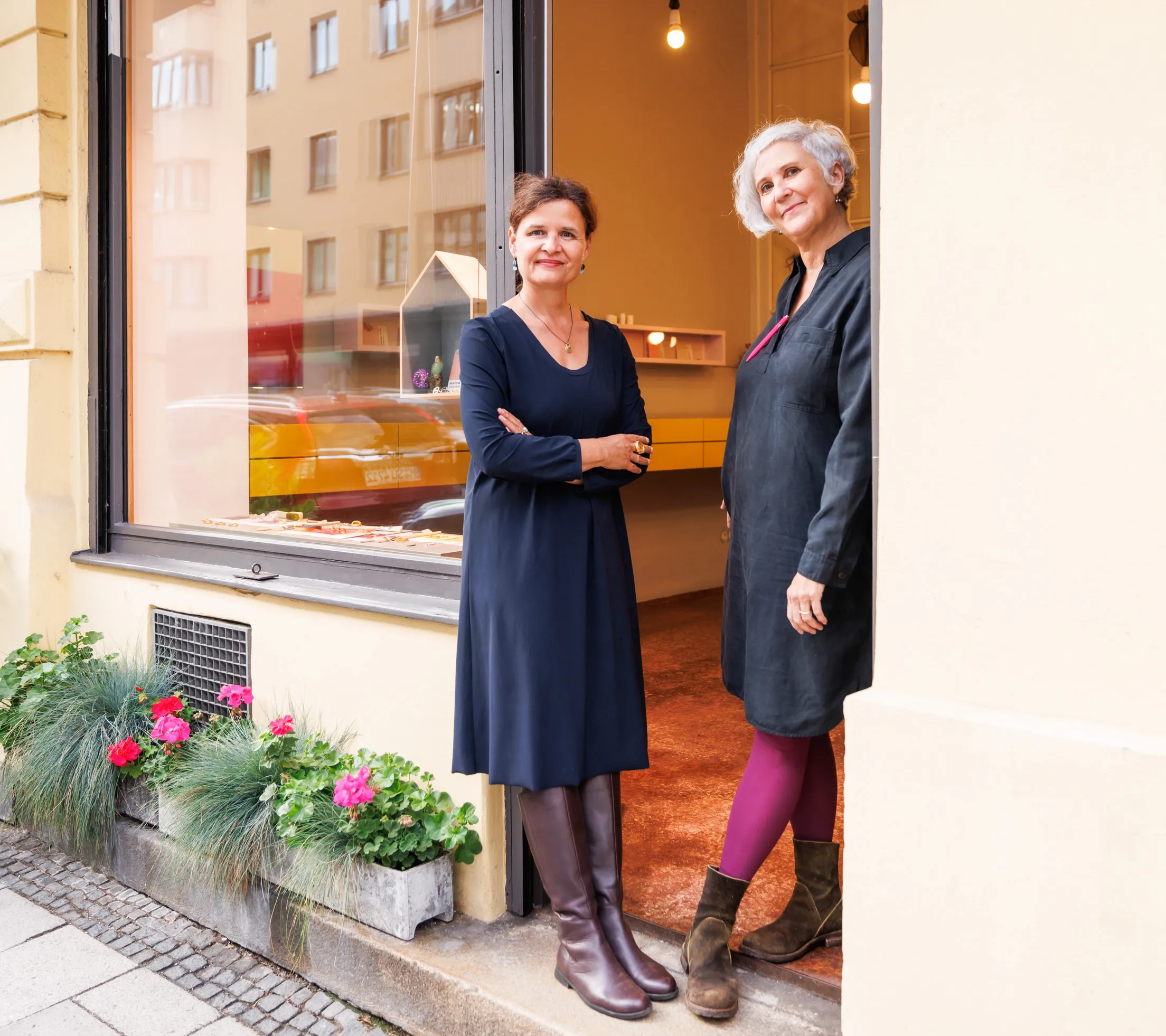 Zwei Inhaberinnen eines Schmuckladens stehen entspannt lächelnd in der Eingangstür, links die Dame im blauen Kleid hat die Arme verschränkt und rechts steht eine Dame im schwarzen Kleid mit lila Strumpfhose an die Wand gelehnt. Links befindet sich ein Schaufenster mit einer Schmuckauslage und auf dem Steinboden stehen zwei Blumenkästen.