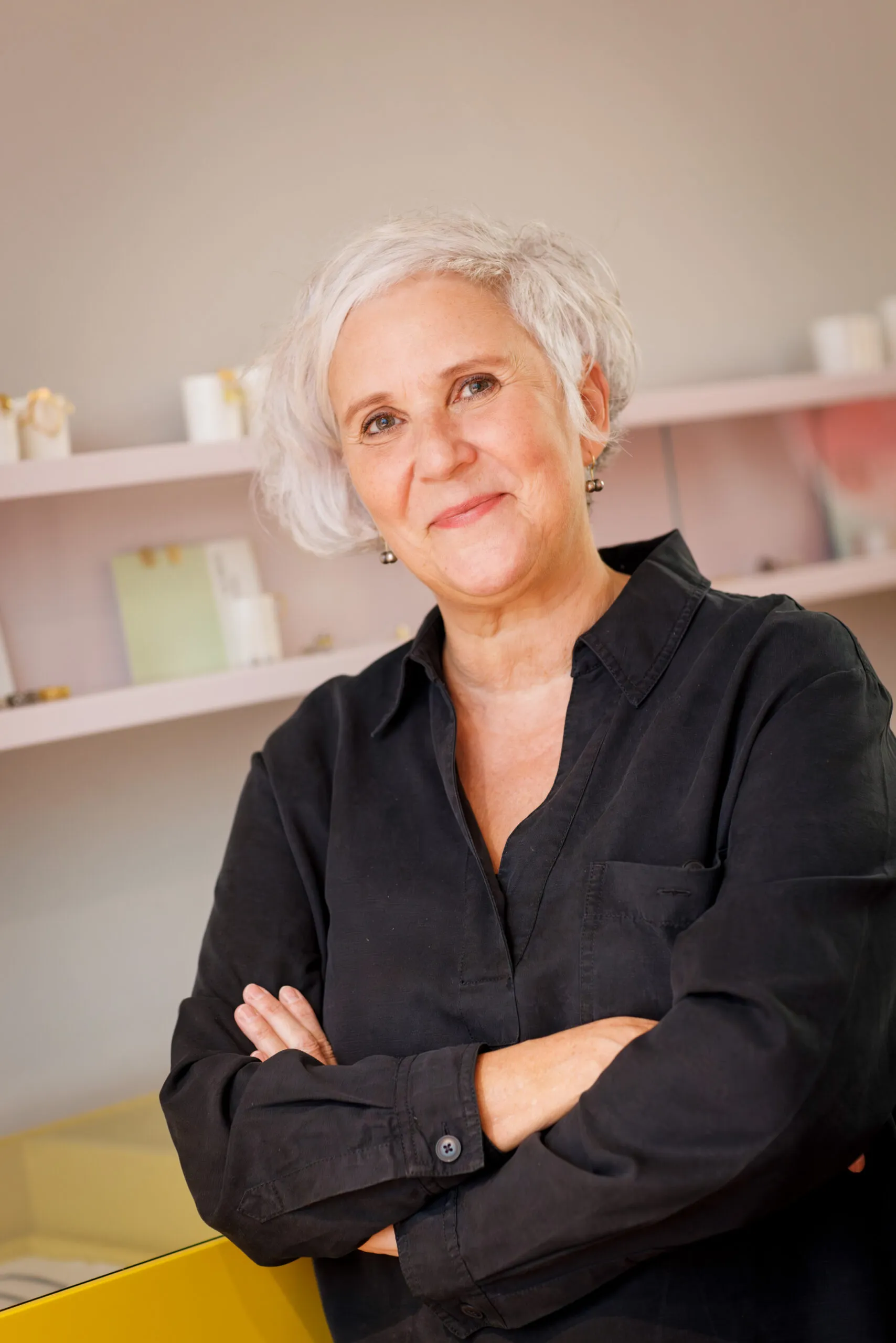 Portraits einer Goldschmiedin, sie trägt ein schwarzes Kleid, hat die Arme verschränkt und schaut in die Kamera. Sie hat graues, kurzes Haar und Ohrringe. Im Hintergrund unscharf ist eine helle Wand, mit zwei Wandregalen und ein Stück einer gelben Vitrine zu sehen.