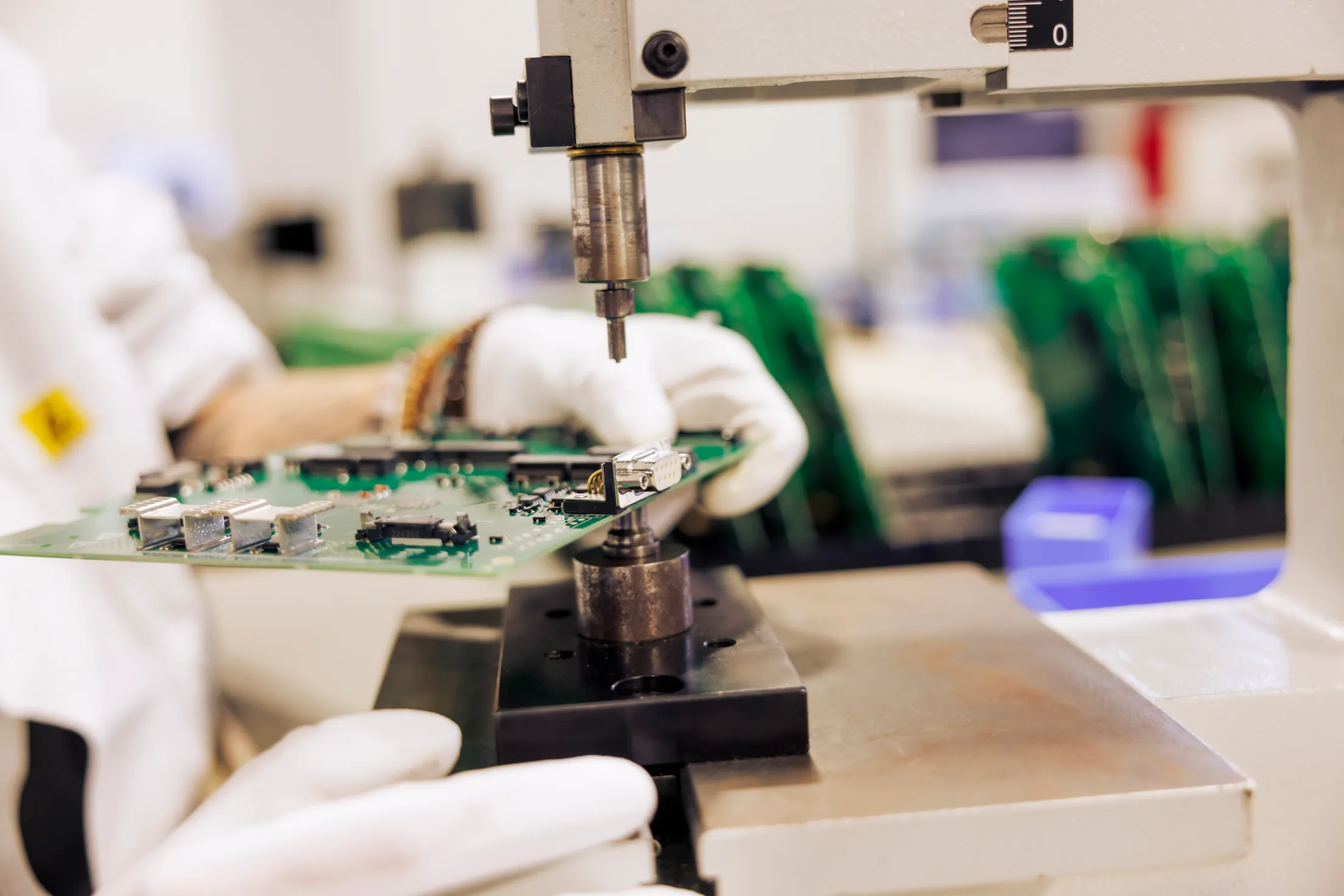 Closeup von zwei Händen mit weißen Handschuhen, die eine Platine halten. Diese wird von einer Maschine bearbeitet.