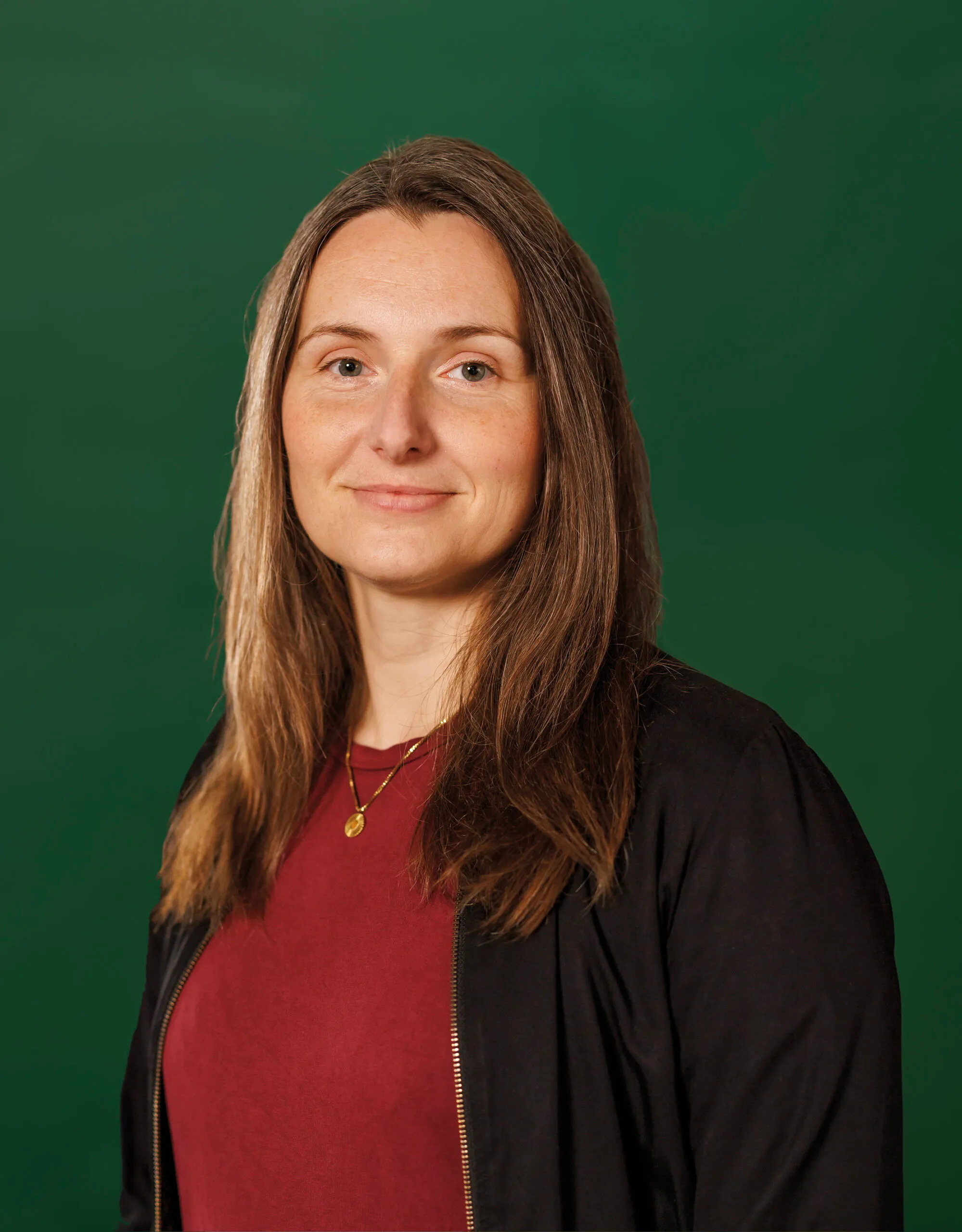 Frau mit braunen langen Haaren schaut selbstbewusst in die Kamera. Sie trägt ein rotes Shirt und eine schwarze Jacke, Hintergrund ist dunkelgrün.