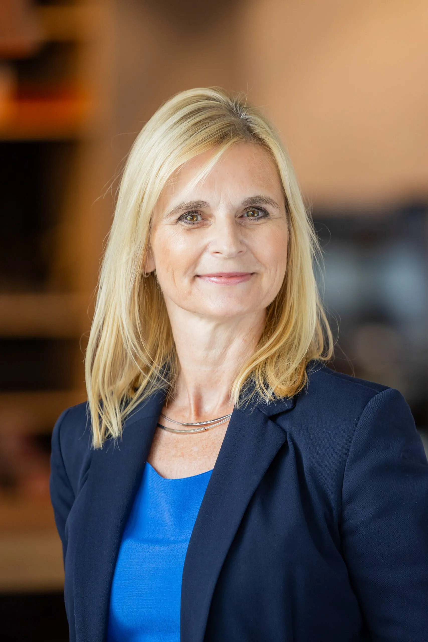 Portraits einer blonden Dame, mit mittellangen, glattem Haar. Sie steht leicht gedreht und lächelt in die Kamera. Sie trägt ein dunkelblaues Jacket und darunter ein königsblaues Shirt. Der Hintergrund ist orange und unscharf, links sind unscharf dunkle Regale zu erkennen.