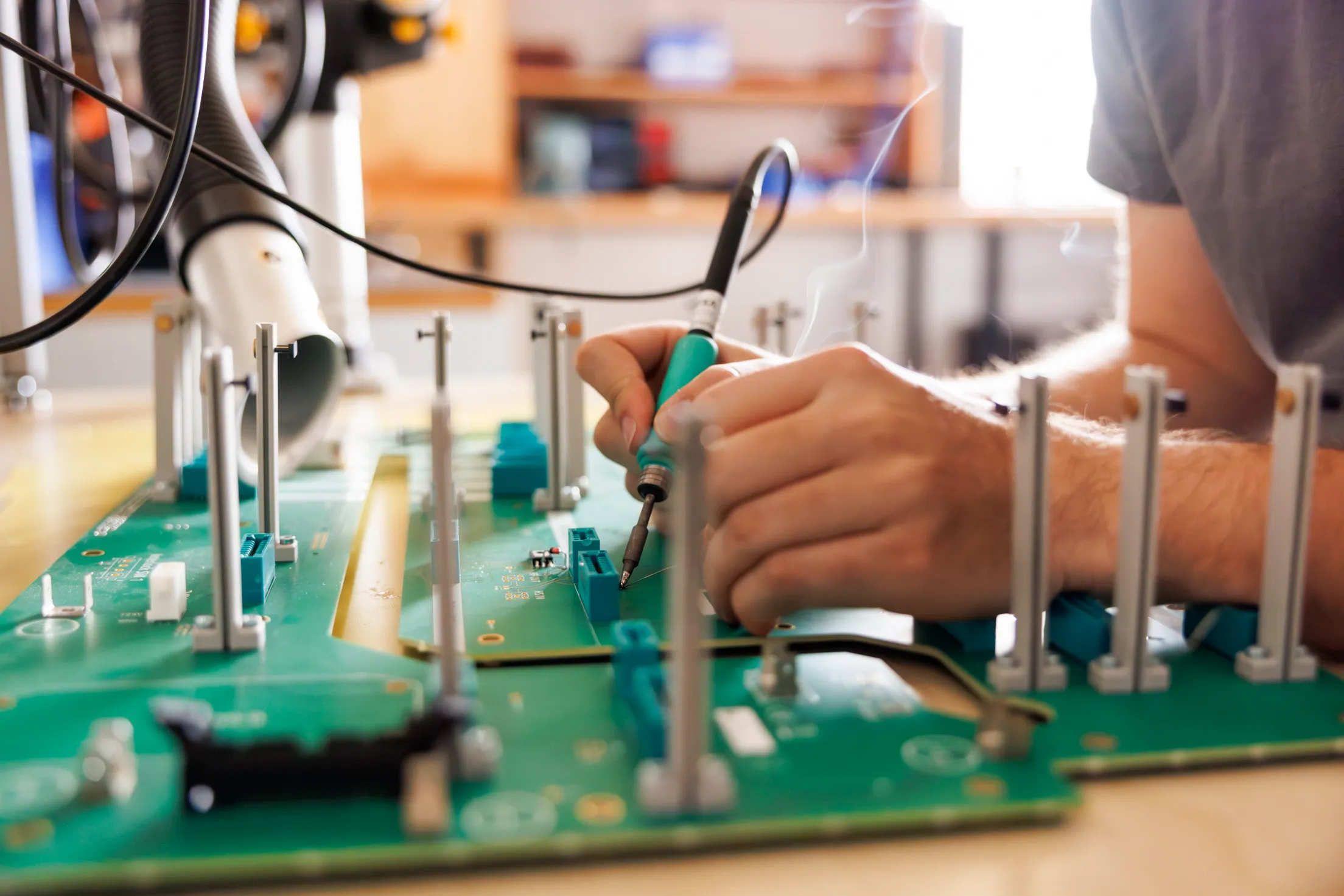 Zwei Hände sind zu sehen, die rechte Hand hält ein Lötkolben und lötet etwas auf einer grünen Platine. Rauch ist zu sehen und das Bild ist in warmen Tönen gehalten.
