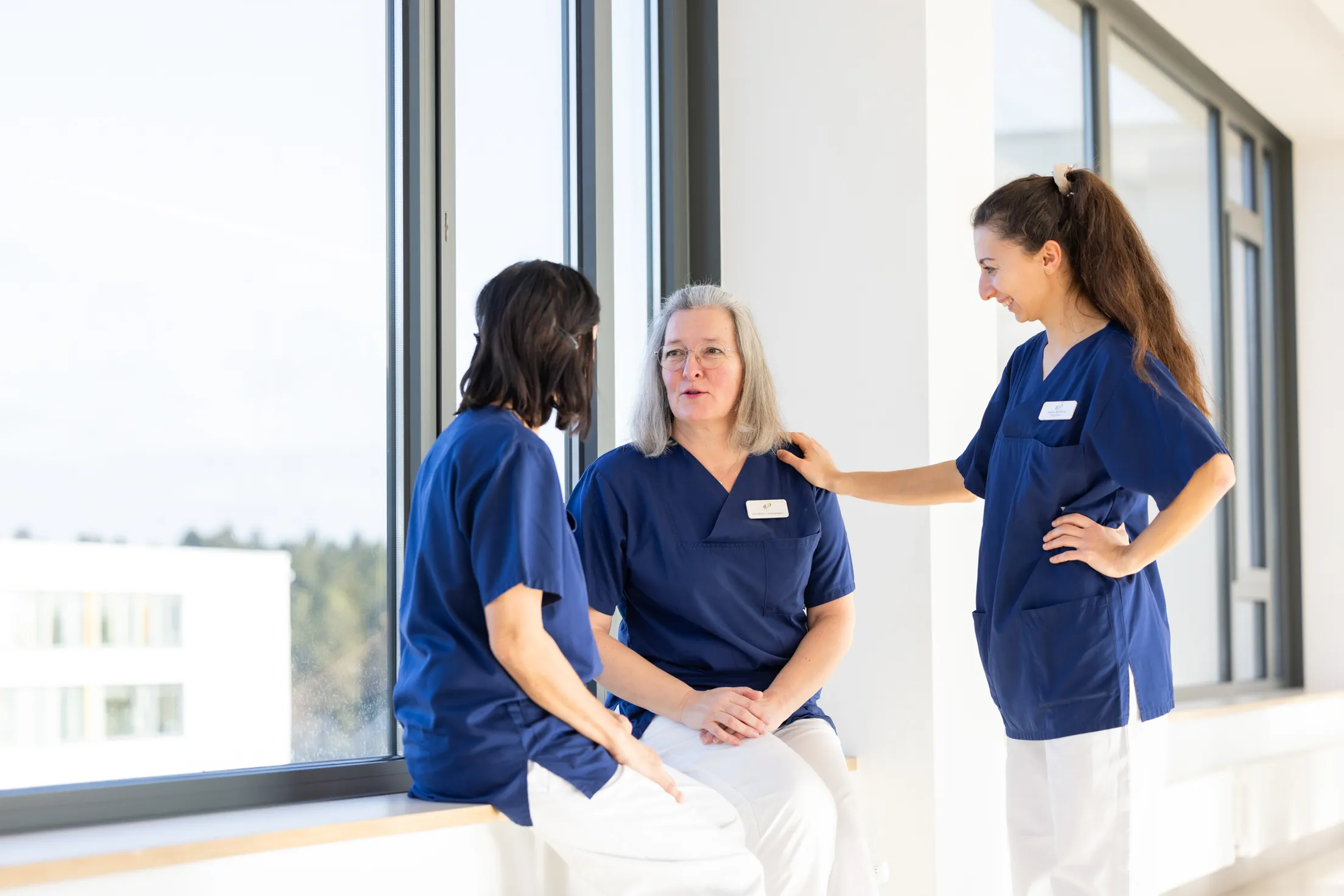 Drei Pflegekräfte sind in einer Gruppe zusammen am Fenster und unterhalten sich in einer lockeren Atmosphäre. Alle drei tragen Klinikkleidung: dunkelblaue Kasack und weiße Hosen. Zwei Damen sitzen auf einer Fensterbank, links die Frau mit schwarzen halblangen Haaren mit dem Rücken zum Betrachter, in der Mitte ein etwas ältere Dame mit grauen schulterlangen Haaren und einer hellen Brille, rechts im Foto steht eine jüngere Schwester mit dunkelbraunen sehr langen Haaren zu einem Zopf gebunden. Sie hat ihre rechte Hand auf der Schulter der älteren Kollegin in der Mitte. Der Fenstergang ist hauptsächlich weiß, nur die Fensterrahmen sind dunkelgrau.