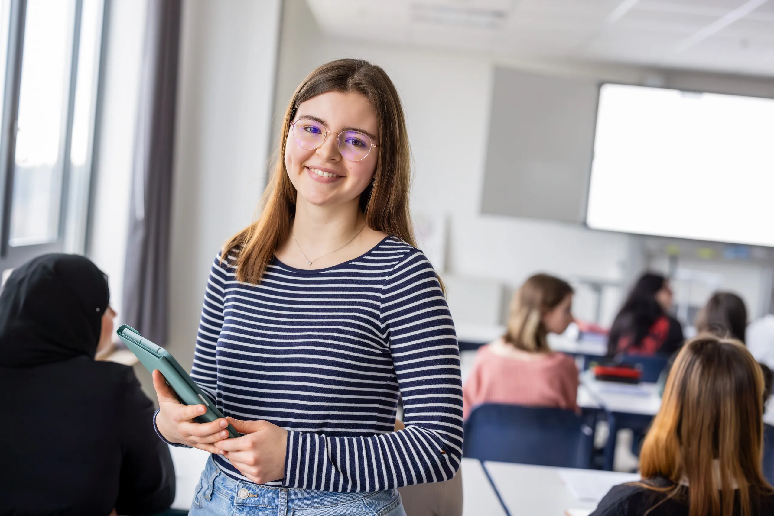 Ein Klassenzimmer der Pflegeschule ist auf dem Foto zu sehen mit mehreren Schüler /- innen unscharf von hinten, ein heller großer Monitor in der Mitte, links eine Fensterreihe. Links die Schülerin trägt eine schwarze Kopfbekleidung. Daneben steht eine junge Schülerin mit dem Körper zur Kamera gedreht. Sie hat dunkelblonde lange Haare, eine goldene Brille und ein langärmeliges, blau-weiß gestreiftes Shirt an. Sie hält auf ihre rechte Seite gedreht ein blaugrünes Tablet in der Hand und lächelt in die Kamera.