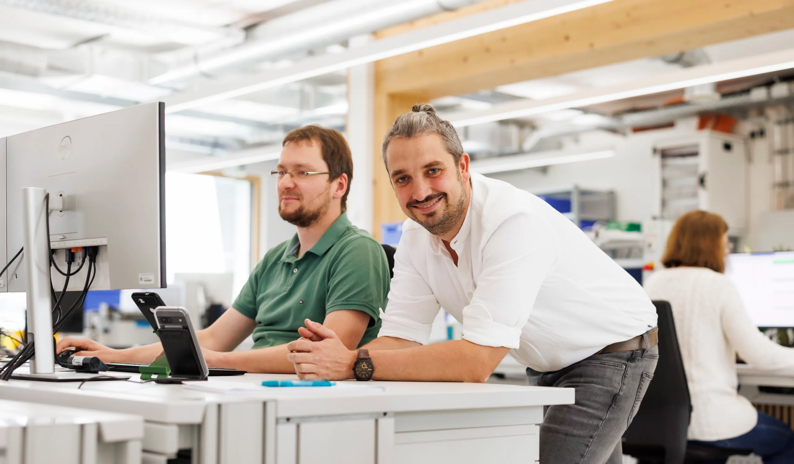 Drei Personen sind in einem hellen Großraumbüro zu sehen. Ein Mann mit weißem Hemd und cooler Frisur lächelt selbstbewusst in die Kamera. Er stützt sich lässig auf einen Tisch. Die anderen beiden Personen arbeiten an Computern und sind unscharf fotografiert.