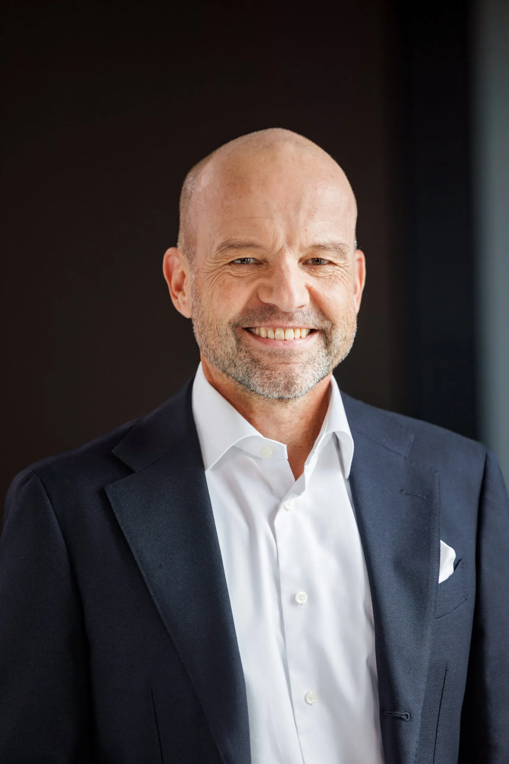 Portraits von der Geschäftsführung, ein Mann mittleren Alters, mit Glatze und grauem Bart. Er trägt ein weißes Hemd, ein dunkelblaues Sakko mit weißem Einstecktuch und lächelt in die Kamera. Im Hintergrund dunkle, unscharfe Strukturen in Schwarz und Grautönen.