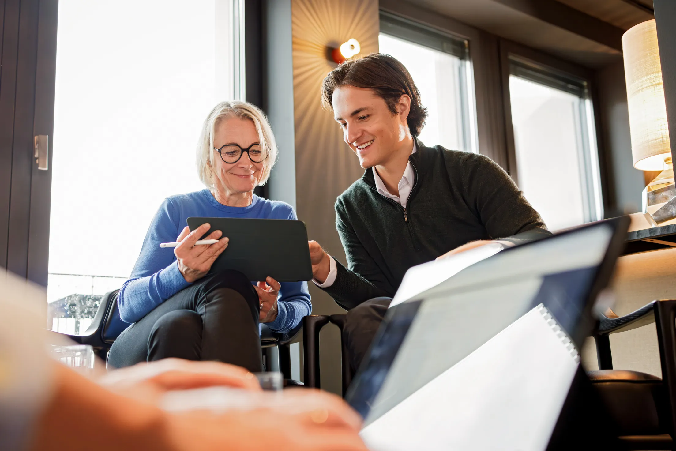 Zwei Personen sitzen auf Stühlen an einer Fensterreihe mit grauem Rahmen und Säule und schauen in ein Tablet zusammen. Links eine Frau mit blondem kinnlangem Haar in einem blauen Pullover und schwarzer Brille hält das Tablet und einen weißen digitalen Stift. Rechts schaut ein jüngerer Mann mit braunem Haar in einem dunkelgrünen Pullover in das Tablet. Im Vordergrund unscharf Hände von einer anderen Person, die an einem Computer arbeiten
