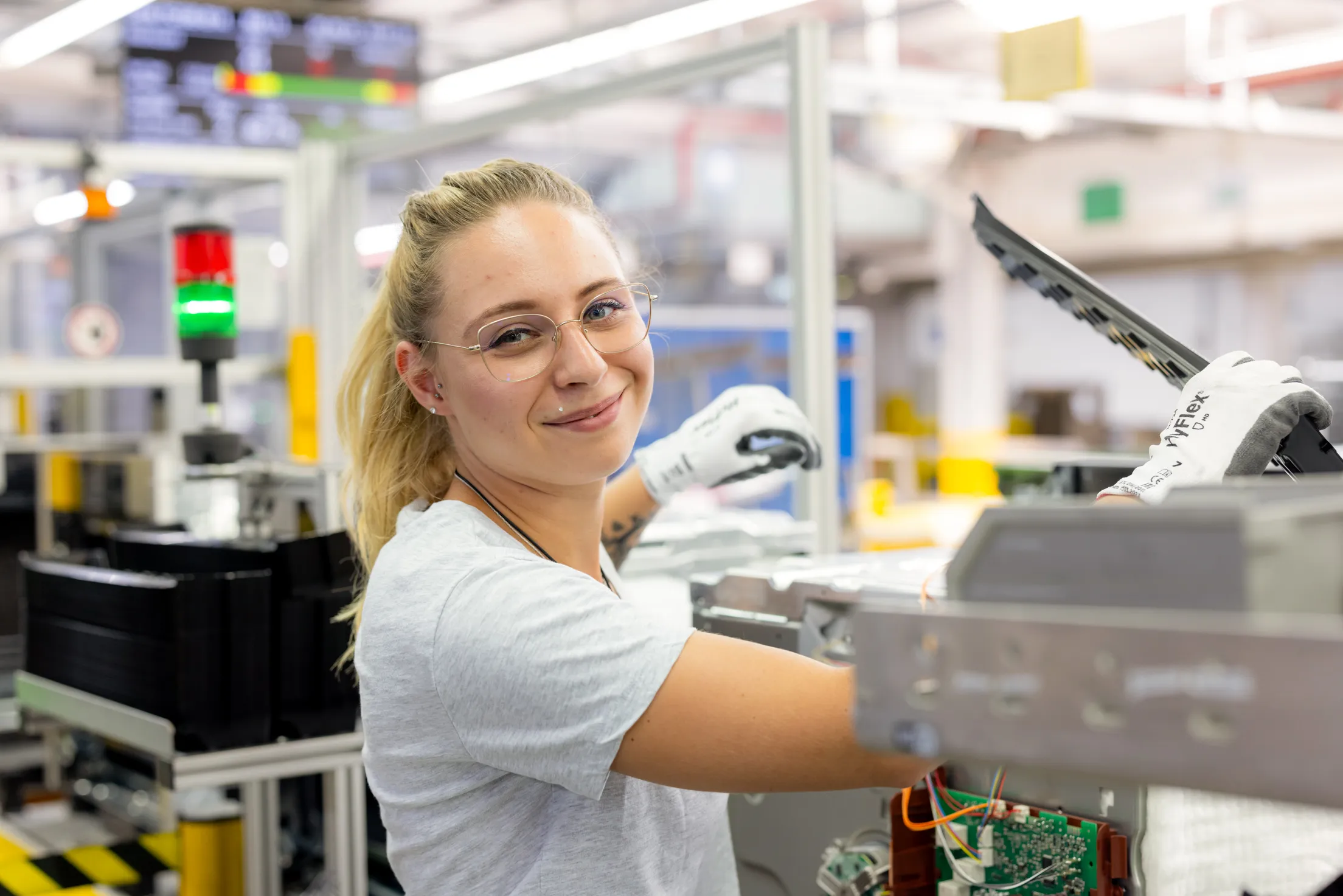 Eine junge Mitarbeiterin mit blondem Zopf, trägt einer Brille, ein Piercing über dem Mund und schaut mit einem sympathischen Lächeln in die Kamera. Sie steht an einer Produktionslinie zwischen Geräten und ist bei der Arbeit, in der rechten Hand hat sein ein Teil des Gerätes in der Hand.