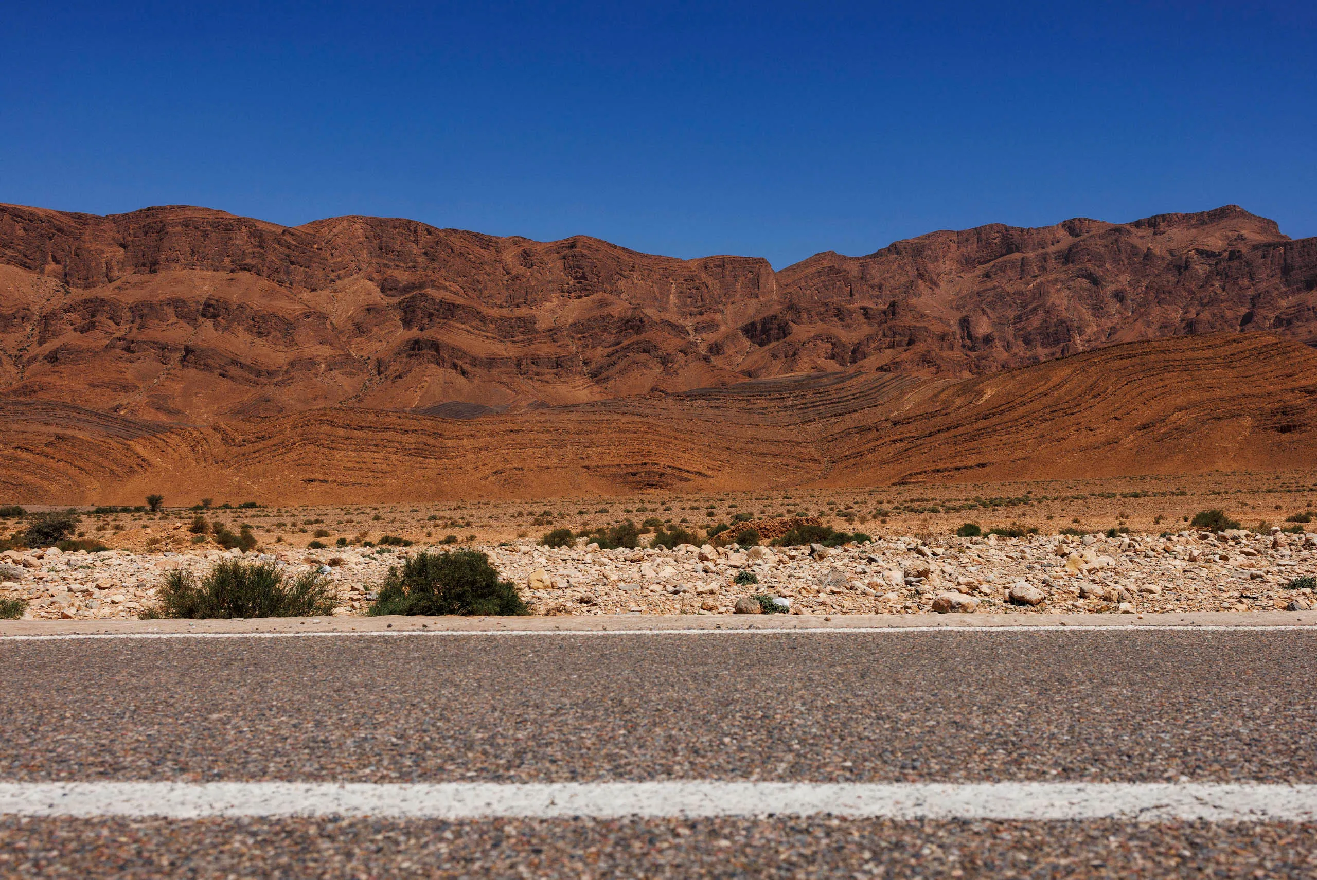 Man sieht eine rötliche Gebirgskette vor blauem Himmel. Darunter sieht man die Steinwüste und die Strasse mit dem weißen Mittelstreifen. Das Bild ist aus der Bodenperspektive aufgenommen.