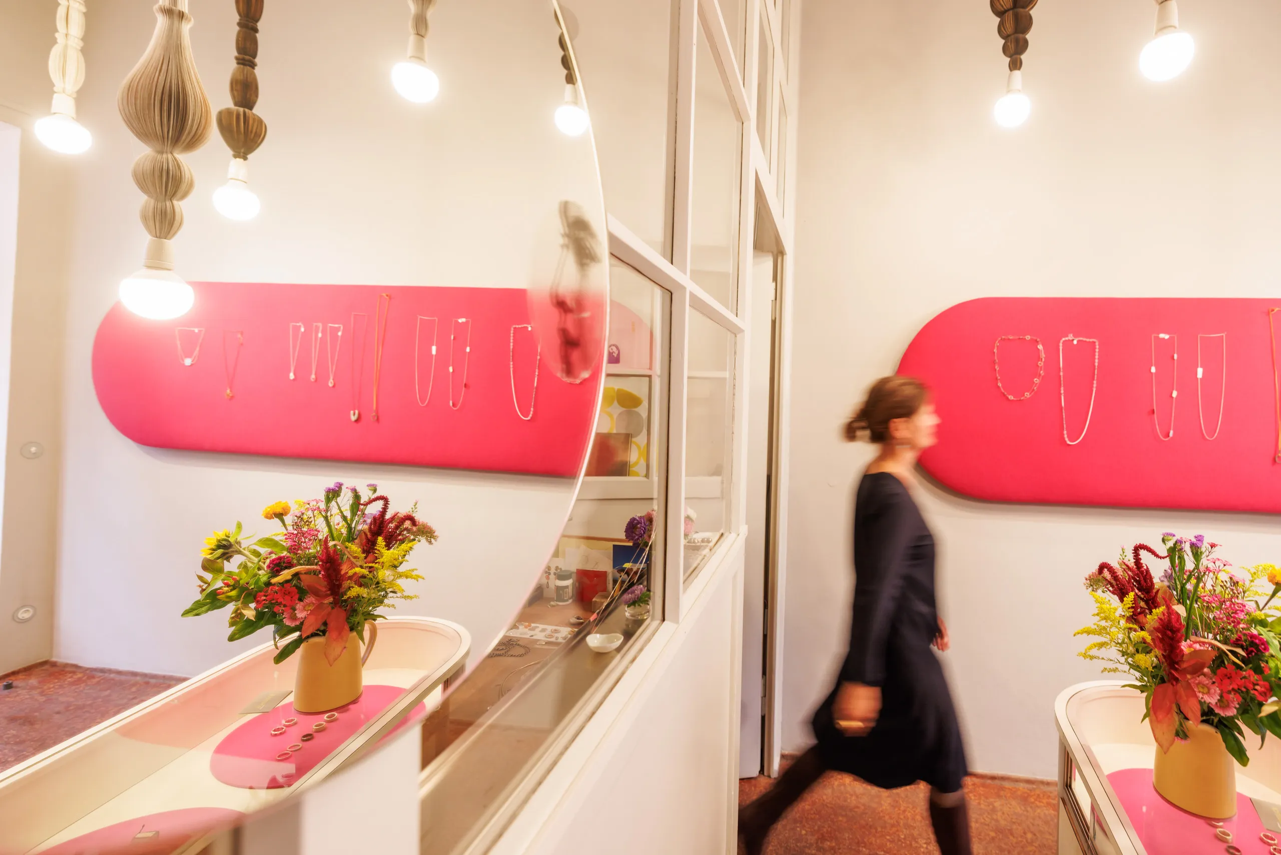 Blick in dem Schmuckladen, links ein Spiegel, in dem sich der Laden spiegelt, Fensterfront und rechts im Anschnitt eine Schmuckvitrine. Auf der Vitrine ein bunter Blumenstrauss. Eine Dame läuft in einem blauen Kleid unscharf von links nach rechts durch das Bild, sie ist verwischt. Im Hintergrund eine rosa Fläche mit vielen Ketten.