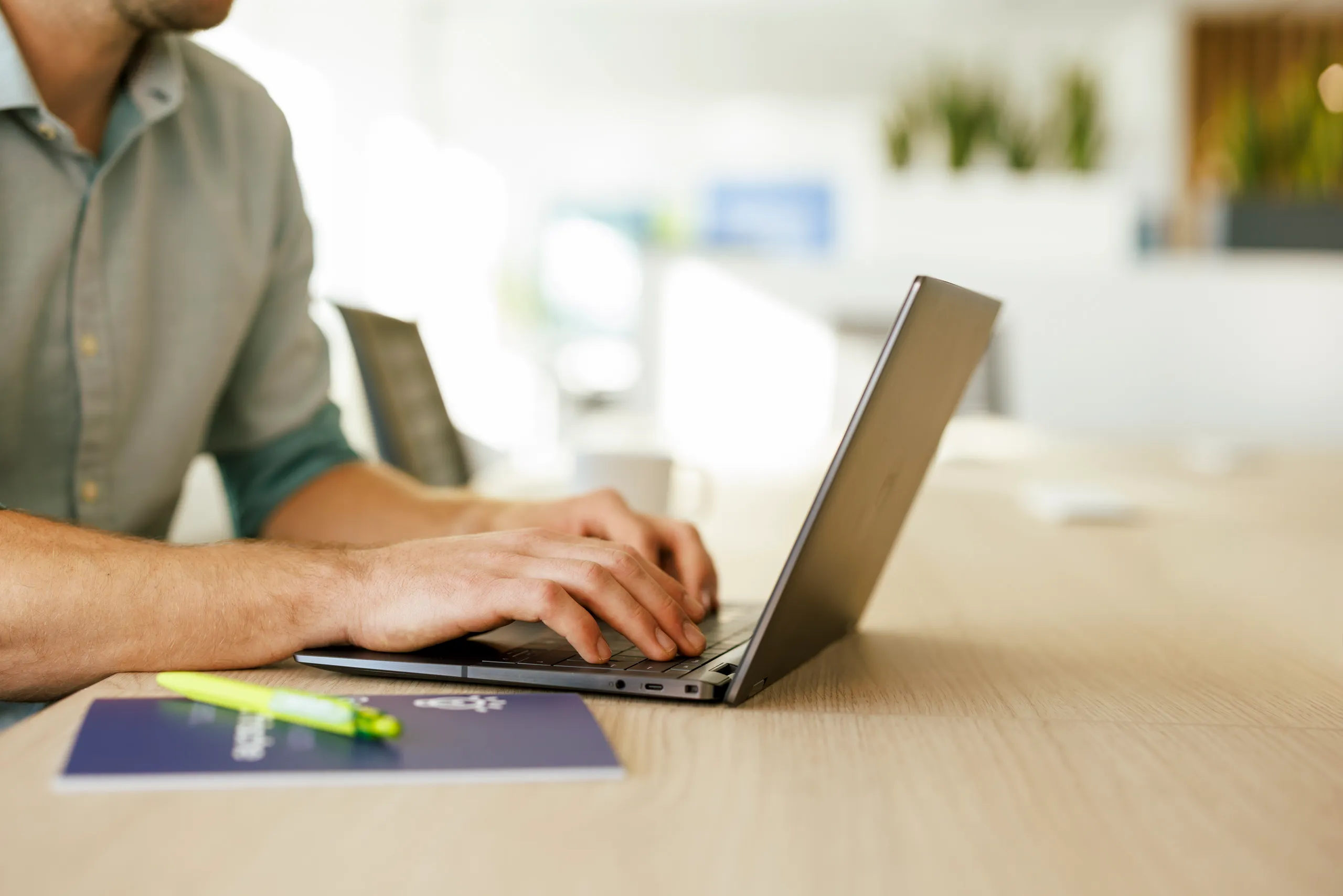 Stillife von einem Laptop mit Händen die tippen, Laptop steht auf einem hellen Tisch, im Hintergrund ist unscharf ein Büro mit schönem Lichteinfall zu sehen
