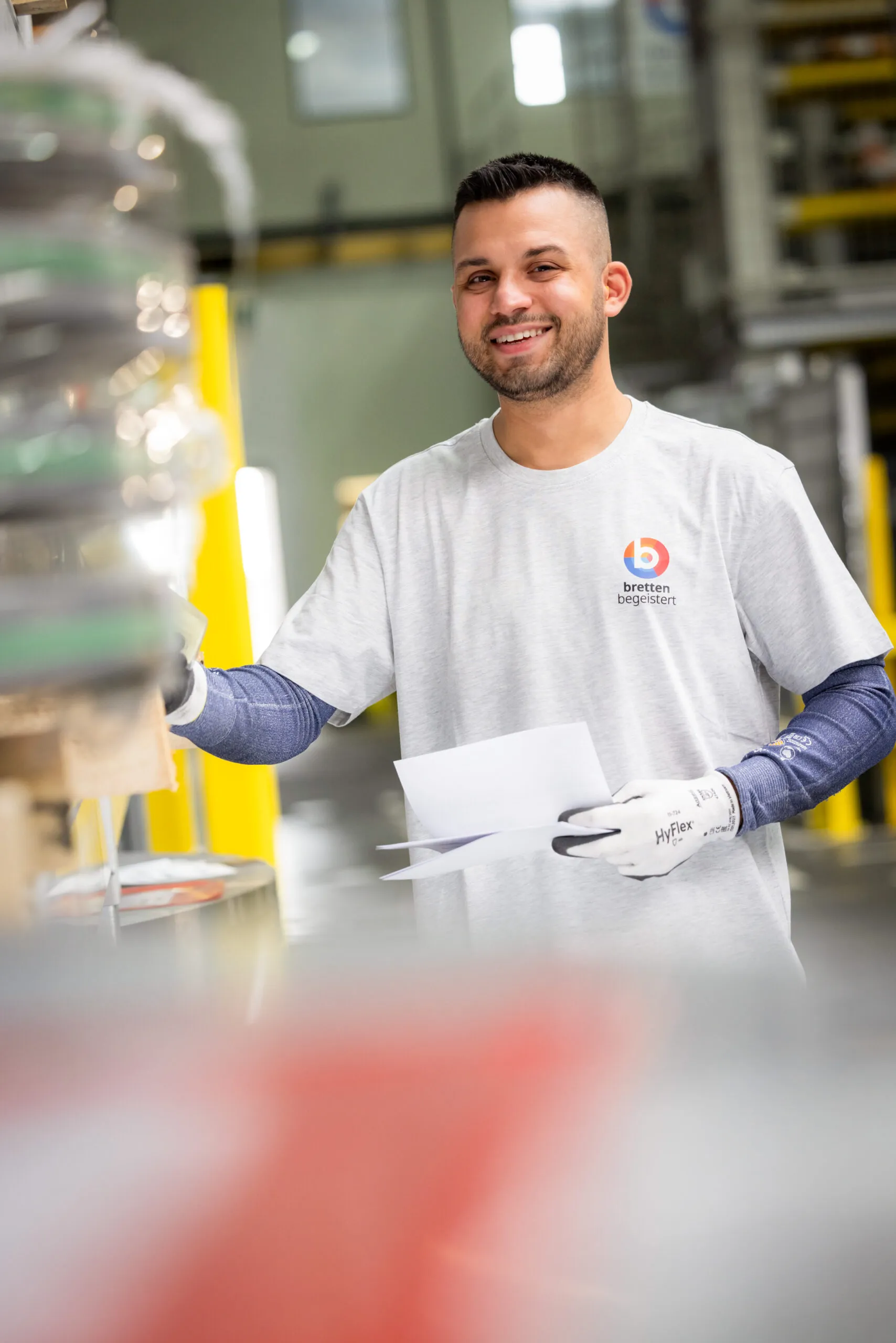 Ein junger Mitarbeiter mit sehr kurzem, dunklen Haar und Bart lacht in die Kamera. Er trägt weiße Sicherheitshandschuhe, blaue Armschoner und ein graues TShirt mit dem Logo „Bretten begeistert“ In seiner rechten Hand hält er ein weißes Blatt Papier. Der Vordergrund unten und an der Seite im Bild ist hell und komplett unscharf.