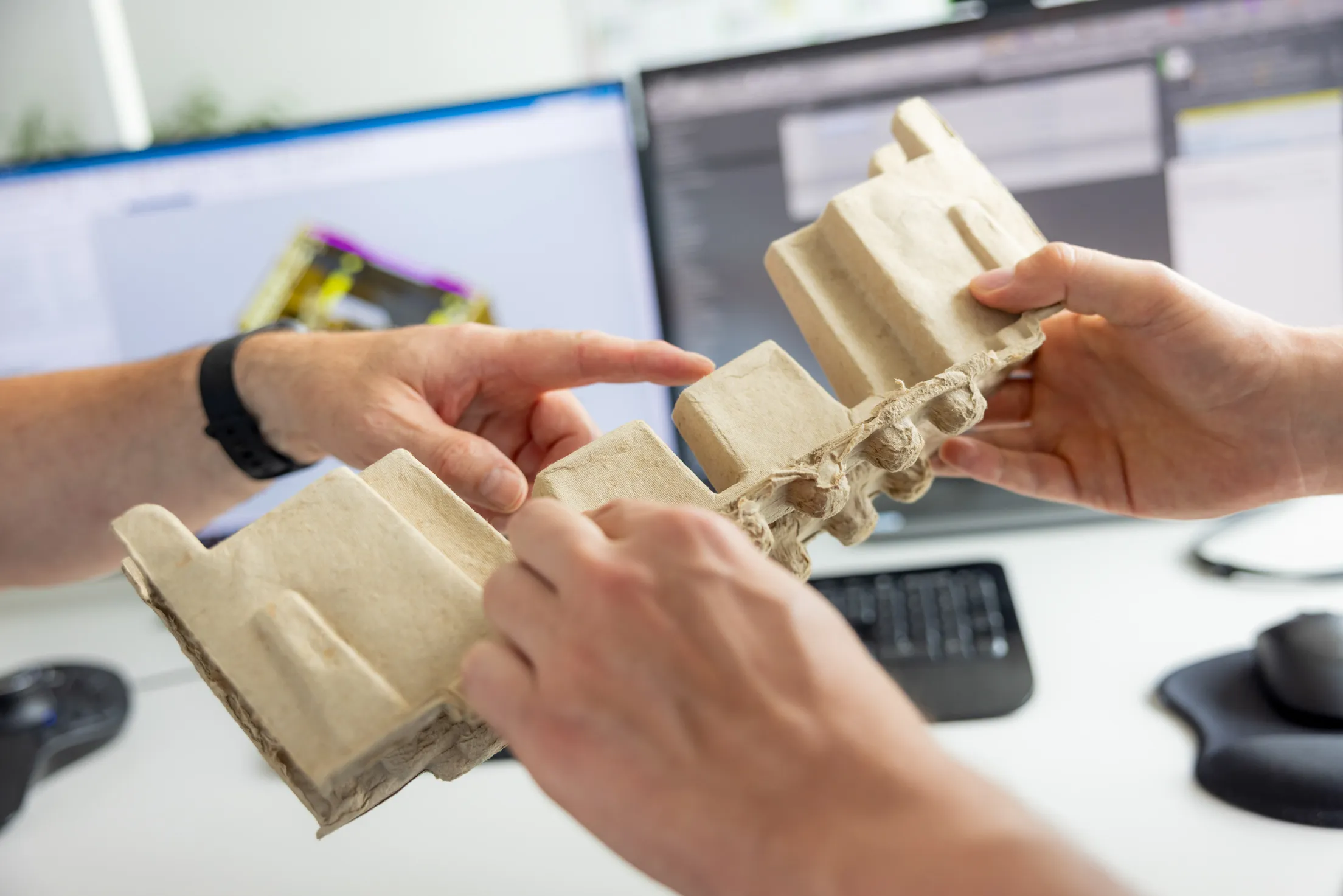 Zwei Hände halten ein Modell aus Pappe in der Hand. Links im Foto befindet sich eine weitere Hand, mit einer schwarzen Armbanduhr und sie deutet mit einem Finger auf das Pappmodell. Im Hintergrund sind unscharf zwei Monitore mit einem 3D Modell auf einem weißen Schreibtisch, eine schwarze Tastatur und eine schwarze Maus abgebildet.