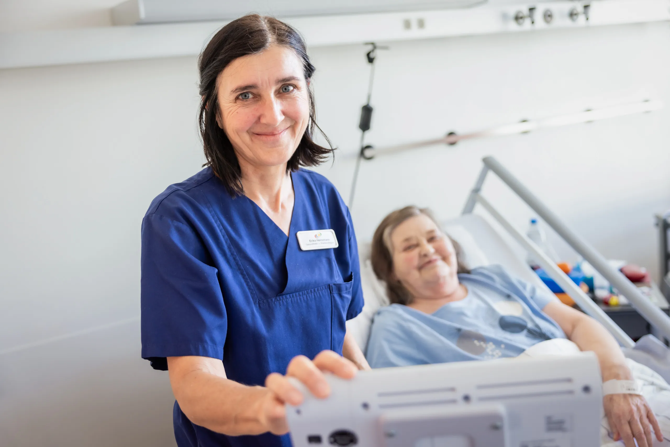 Eine Krankenpflegerin mit dunklen, schulterlangen Haare locker zusammengebunden, ist leicht von oben fotografiert. Sie trägt einen dunkelblauen Kasack, hat ihre rechte Hand an einem Monitor und lächelt in die Kamera. Im Hintergrund unscharf ist ein Patientenzimmer, rechts im Bild einer älteren Dame mit grauen Haaren. Sie liegt in einem Krankenbett mit einem Gitter und trägt ein hellblaues T-Shirt.