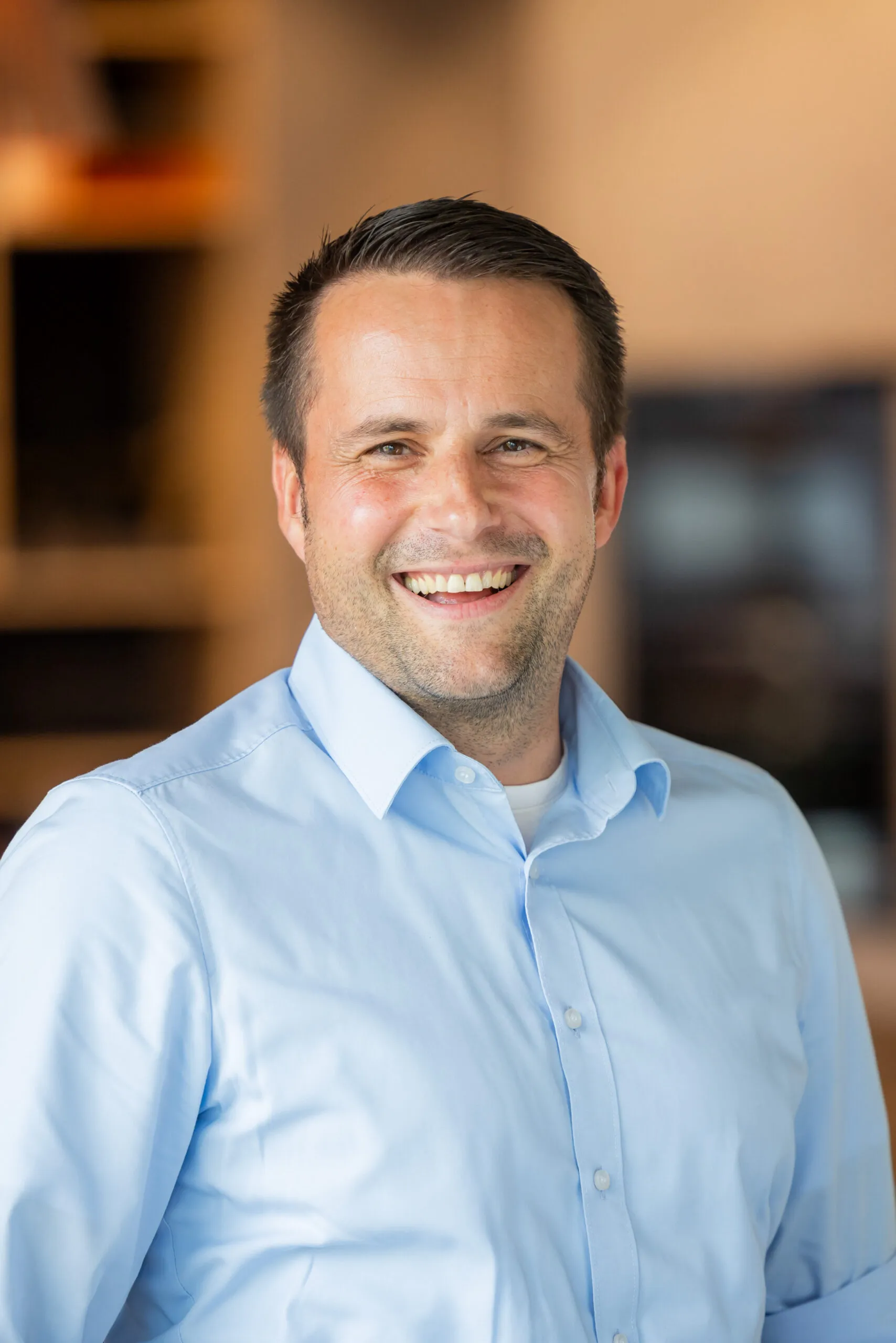 Ein Portraits von einem Mann mittleren Alters, mit braunen kurzen Haaren. Er lacht sehr offen in die Kamera. Er trägt ein hellblaues Hemd mit einem weißen T-Shirt darunter, leicht gedrehte Körperhaltung. Der Hintergrund ist sehr unscharf, in Orangetönen gehalten, links sind unscharf dunkle Regale zu erkennen.