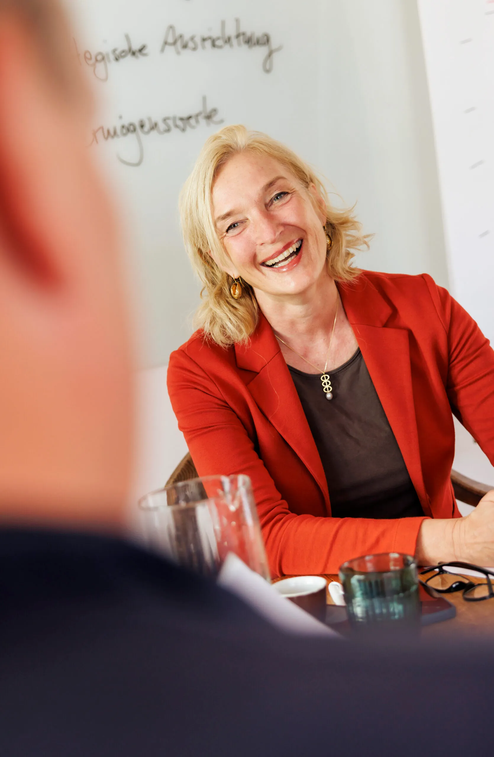 Eine Frau mit rotem Jacket ist im Gespräch mit meinem Mann, der ganz unscharf zu erkennen ist. Die Dame hat eine positive Ausstrahlung.