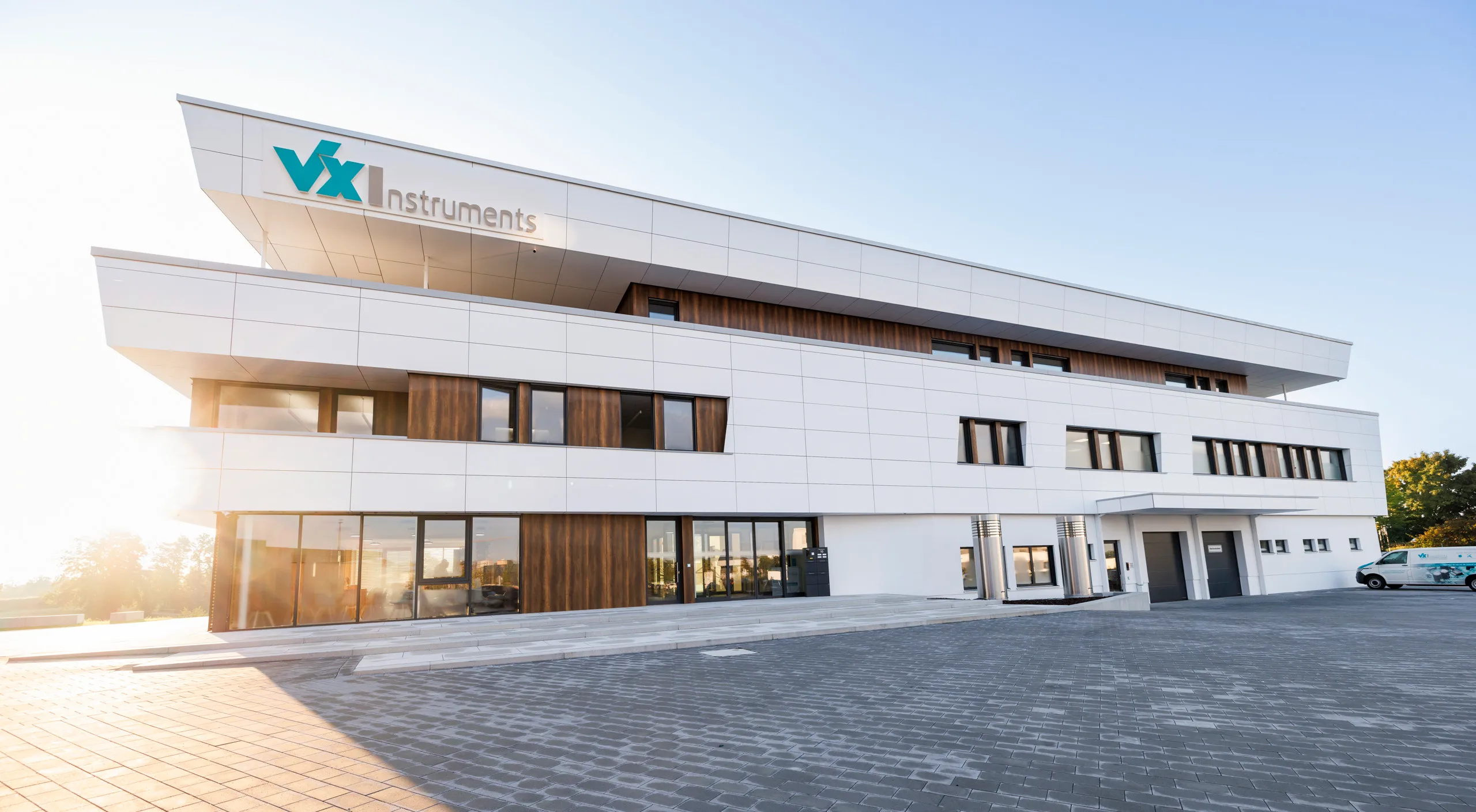 Eine Außenaufnahmen von dem Firmengebäude in der Früh mit der aufgehenden Sonne im Hintergrund ist zu sehen. Das Gebäude ist weiß mit Holzelementen und das Foto ist im Panoramaformat aufgenommen.