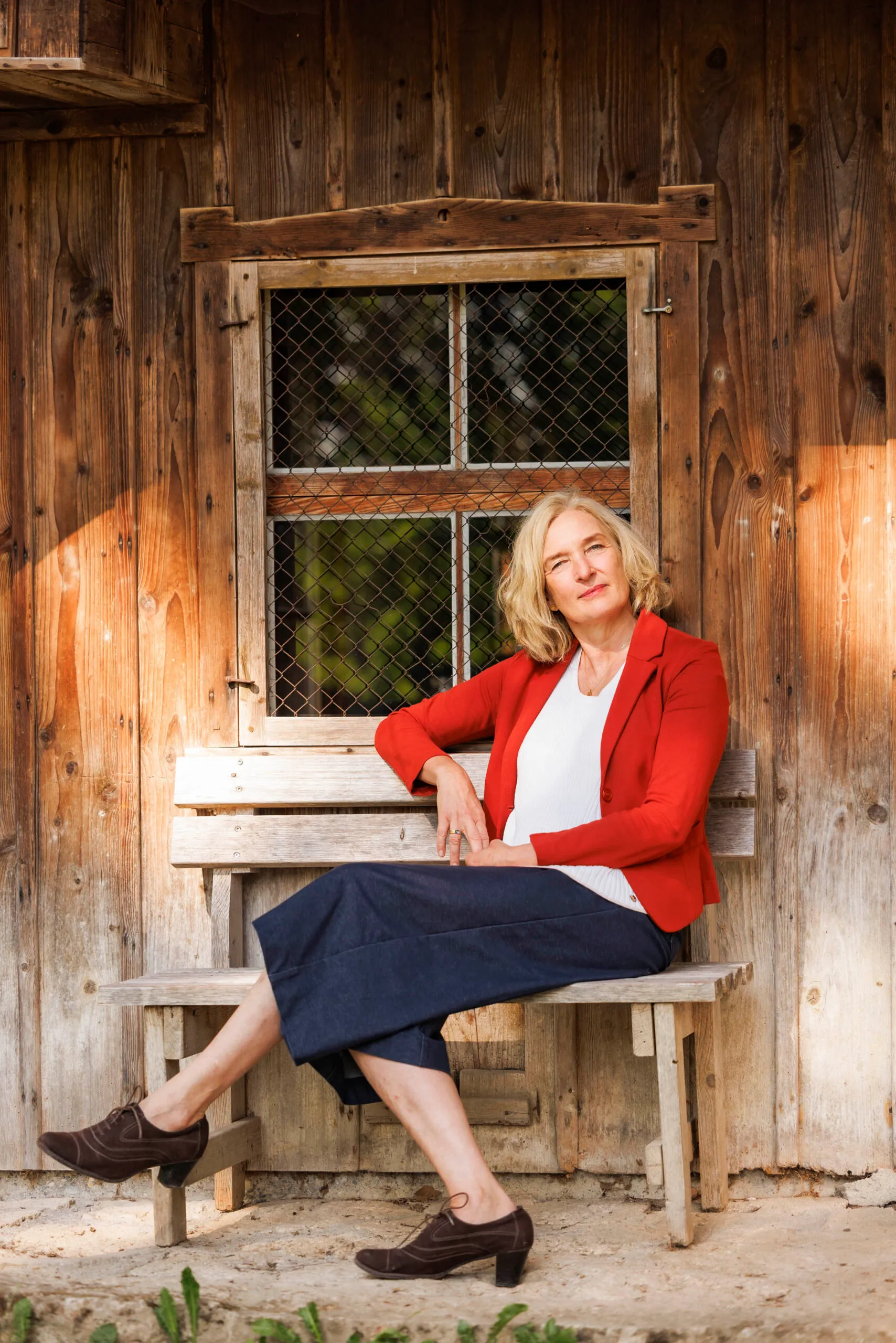 Eine Frau sitzt sehr entspannt auf einer Holzbank vor einer Holzhütte. Sie schaut selbstbewusst in die Kamera und trägt ein knallrotes Jacket und eine blaue 3/4 Hose.