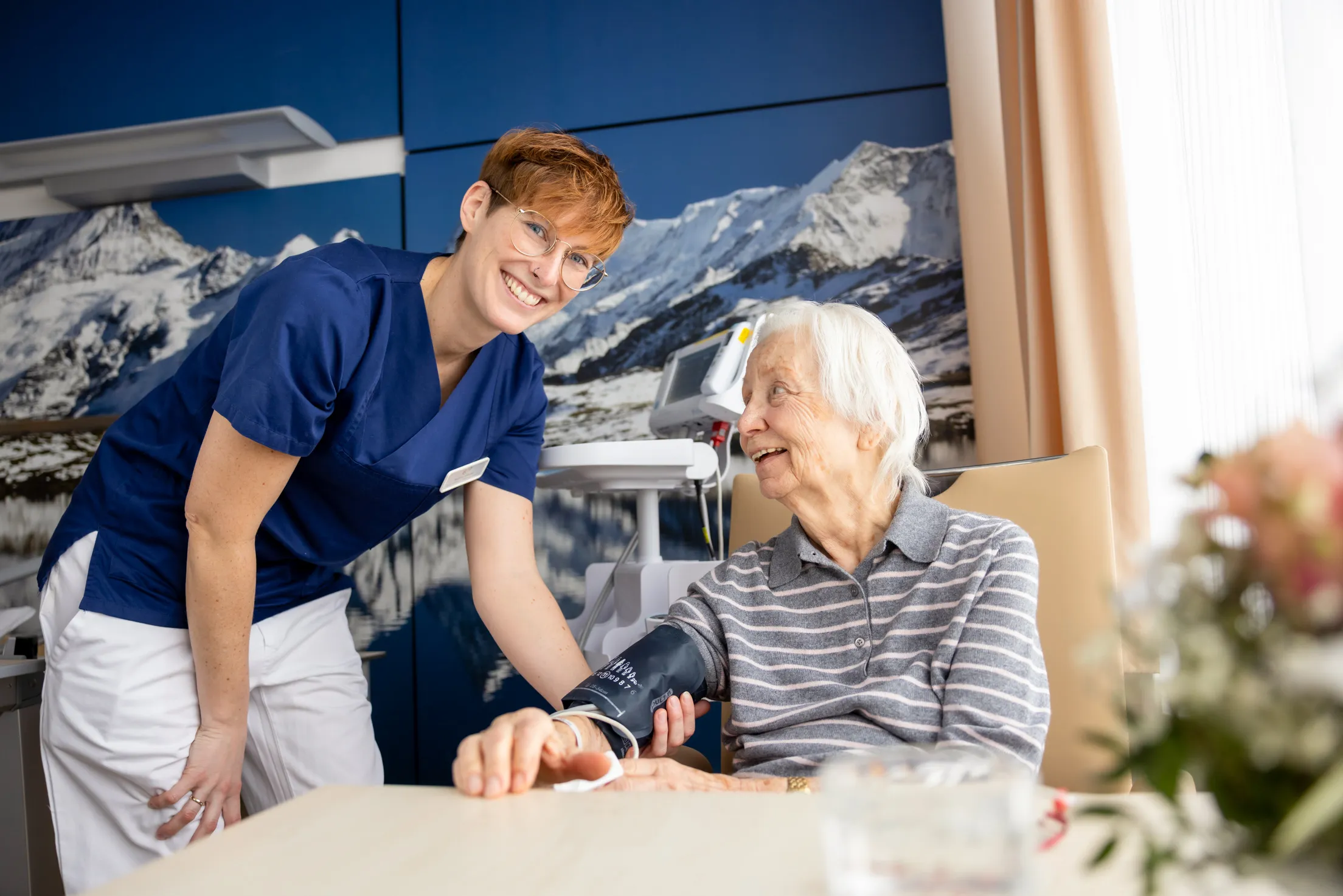Ein Patientenzimmer ist zu sehen mit einer großen Tapete mit Bergpanorama und blauem Himmel im Hintergrund. Rechts hängen beige Vorhänge. Im Vordergrund ist links eine Pflegerin mit kurzem, kupferrotem Haar und einer goldenen Brille. Sie trägt einen dunkelblauen Kasack, eine weiße Hose und beugt sich im Stehen zu einer Patientin, die links von ihr zu sehen ist. Sie misst gerade den Blutdruck und hält den rechten Arm der Patientin. Diese ist im hohen Alter, hat weiße kurze Haare, trägt ein grau gestreiften Pullover und schaut zufrieden zu der Schwester auf.