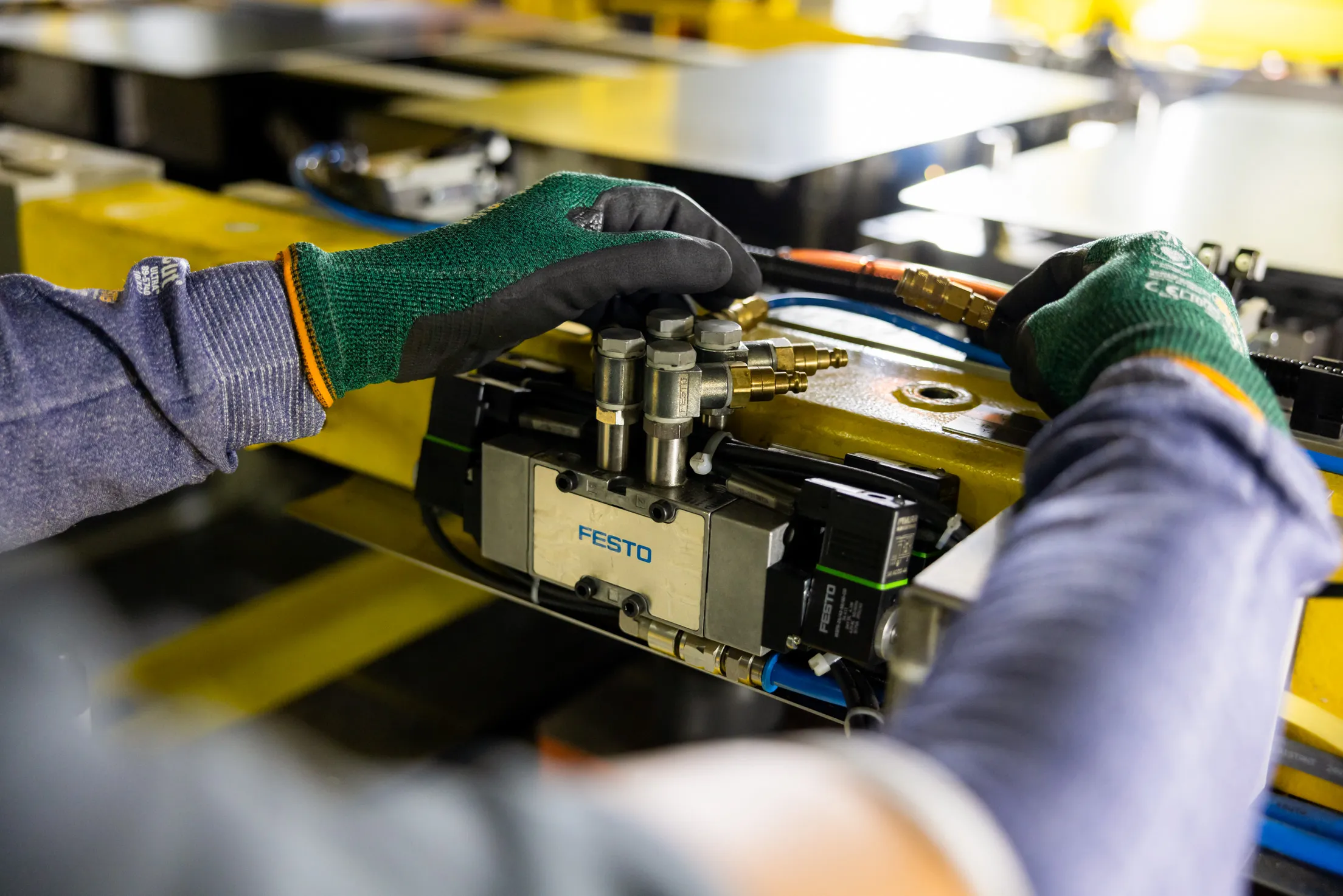 Auf dem Foto ist ein Close-up auf ein Paar Hände mit grün und schwarzen Handschuhen und blauen Armschoner zu sehen. Die Hände sind leicht von oben fotografiert und sie kontrollieren eine Leitung. Das Licht von vorne ist etwas dunkler, dafür viel Gegenlicht von hinten.