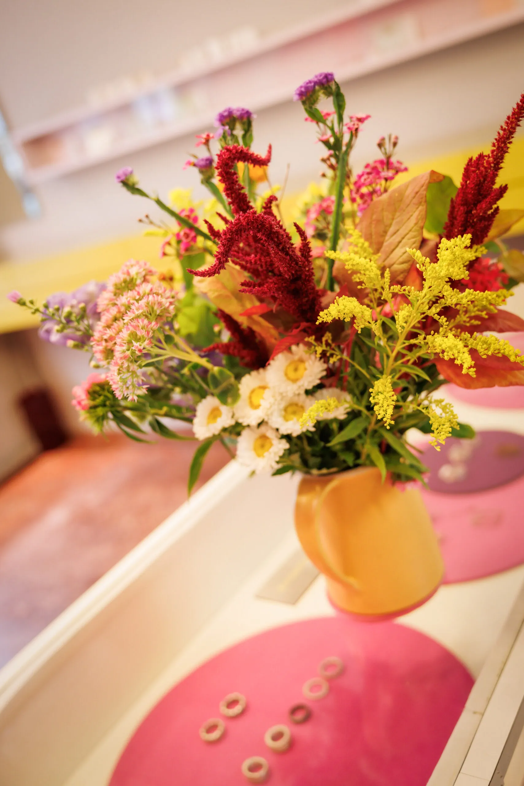 Eine gelbe Blumenvase mit einem bunten Blumenstrauss steht auf einer Schmuckvitrine, die unscharf ist. Ringe liegen auf einem rosa Kreis. Unscharf ist ein Ladenraum mit Regalen an der Wand zu erkennen mit einem rötlichen Boden.