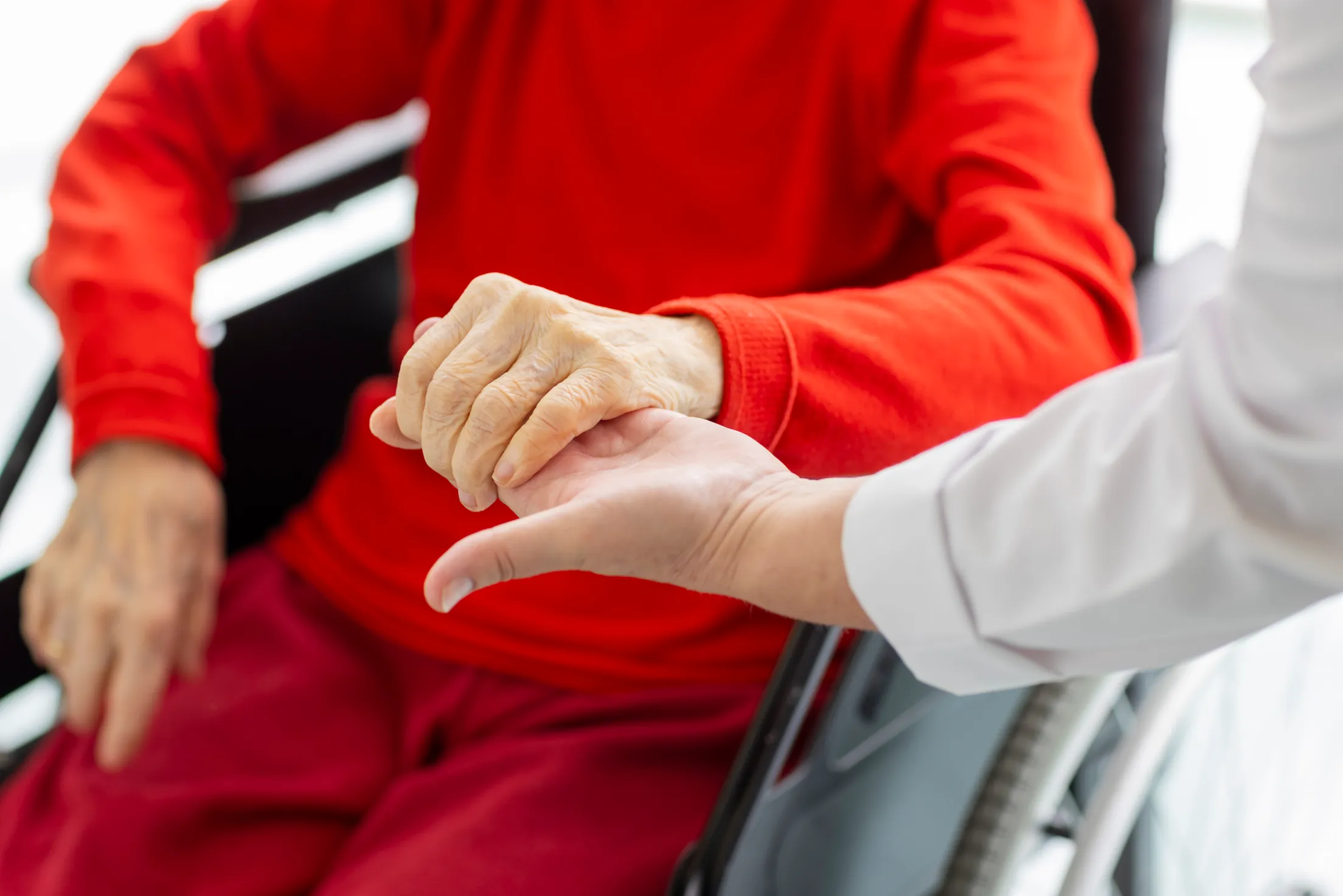 Closeup von zwei Händen, die eine Hand gehört einer Patientin im roter Kleidung und die hält die Hand von einer Dame mit weißem Kittel. Die Patientin sitzt im Rollstuhl.