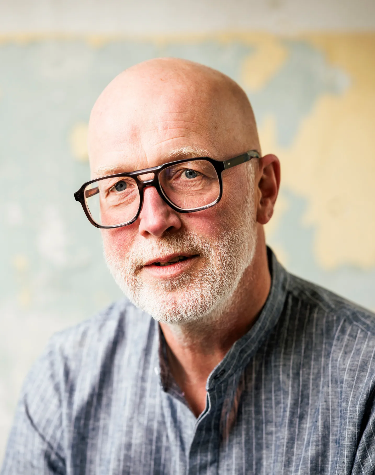 Portraits von einem älteren Mann ohne Haare, auffälliger Brille und einem grauen Bart. Er trägt ein legeres blaues Hemd und schaut direkt in die Kamera