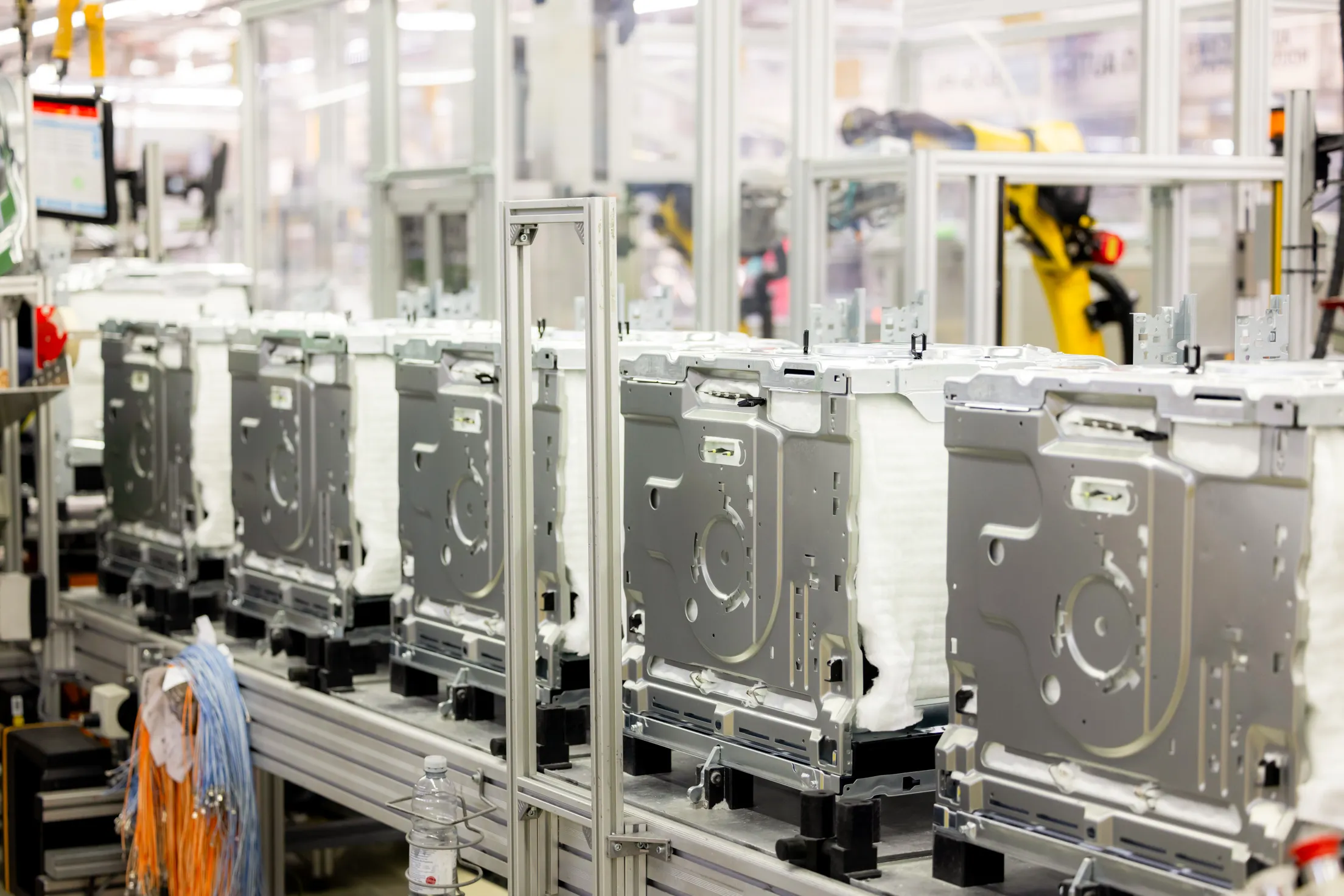 Ein Foto von einem Fließband mit 5 Küchengeräten, Blick auf die silberne Rückseite der Geräte, unscharf im Hintergrund die Fabrikhalle mit Roboter und Maschinen. Links im Bild sind orangefarbene und hellblaue Kabel zu sehen.