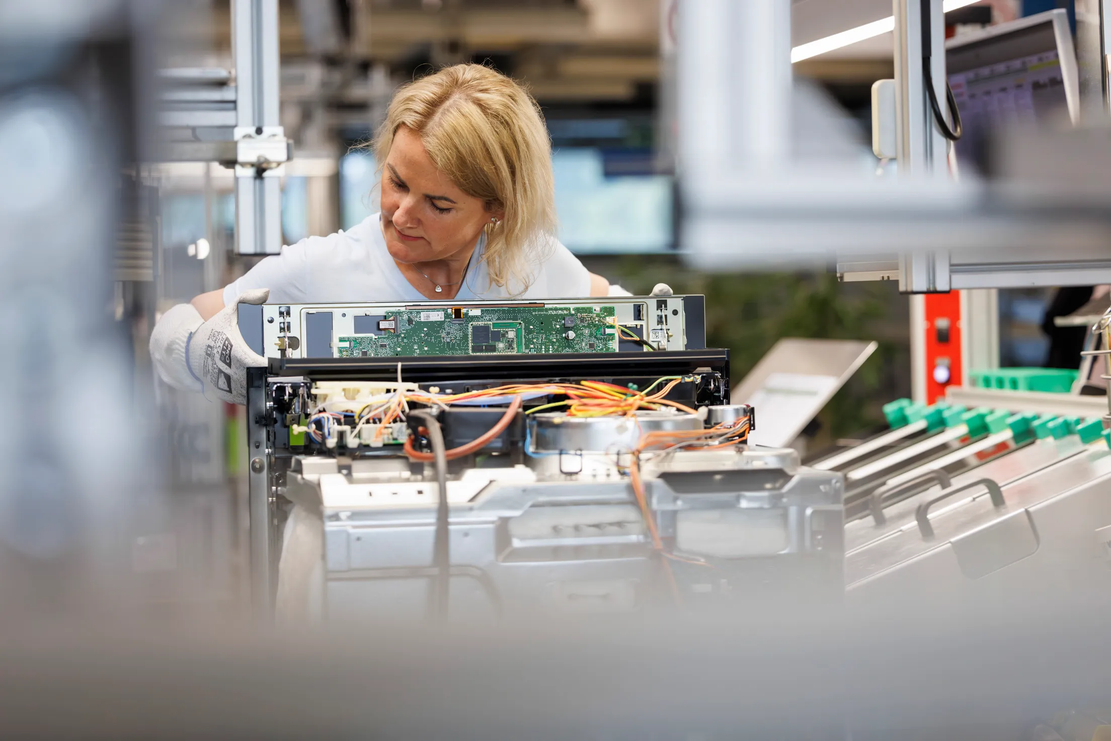 Eine Mitarbeiterin mit hellblauen Shirt, hat blonde kinnlange Haare und steht am Fließband. Sie arbeitet an einem Backofen, dessen Gehäuse hinten offen ist.