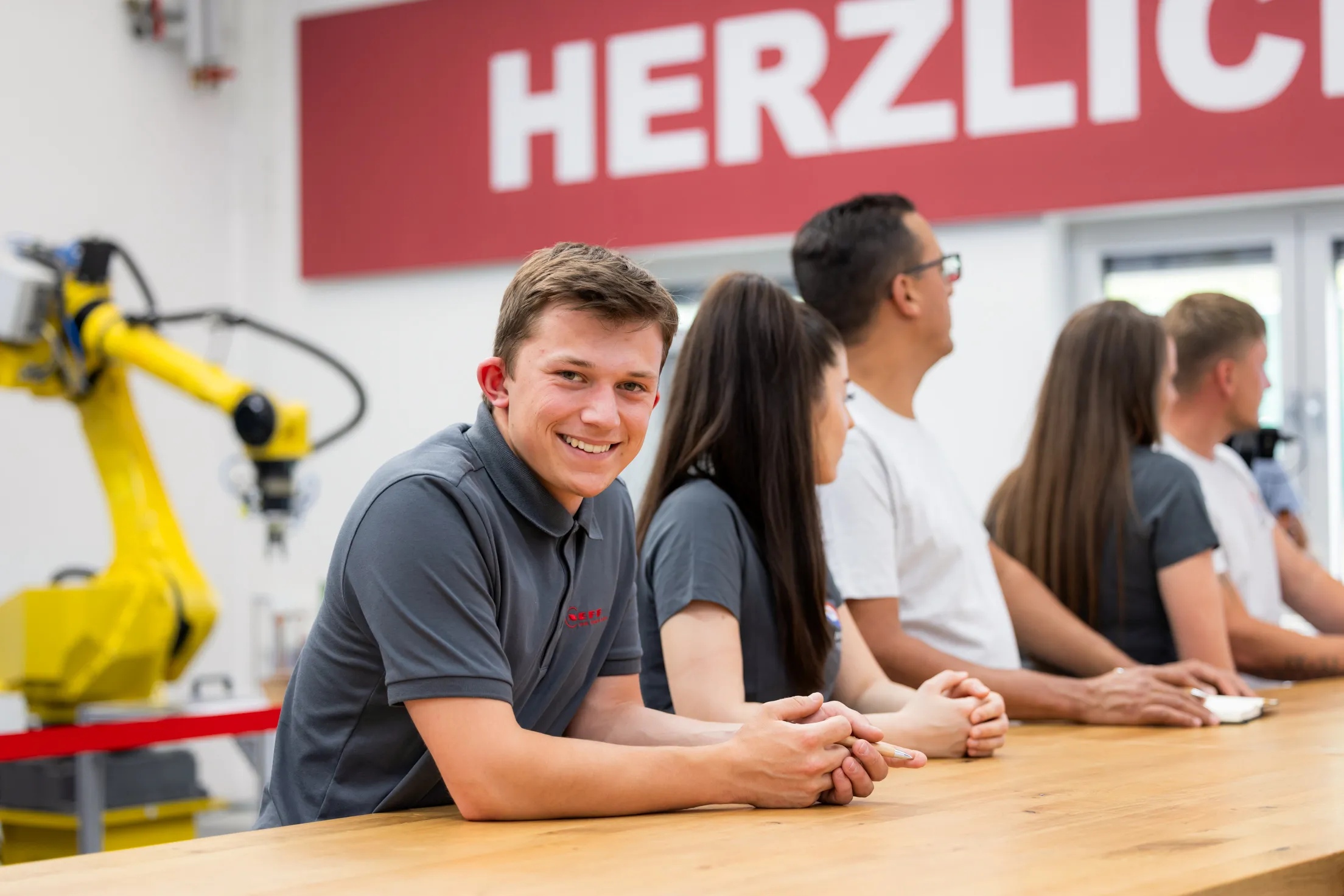 In der Trainingsakademie stehen fünf junge Menschen in einer Reihe an einem langen Holztisch und haben ihre Hände auf den Tisch gelegt. Alle schauen nach rechts, nur ein junger Mann ganz links im Bild schaut direkt in die Kamera und lacht dabei. Er trägt ein graues Poloshirt mit rotem Neff Logo. Links hinten ein gelber Roboter zu sehen und in der Mitte oben ein großes rotes Schild mit weißer Schrift. Herzlich steht darauf.