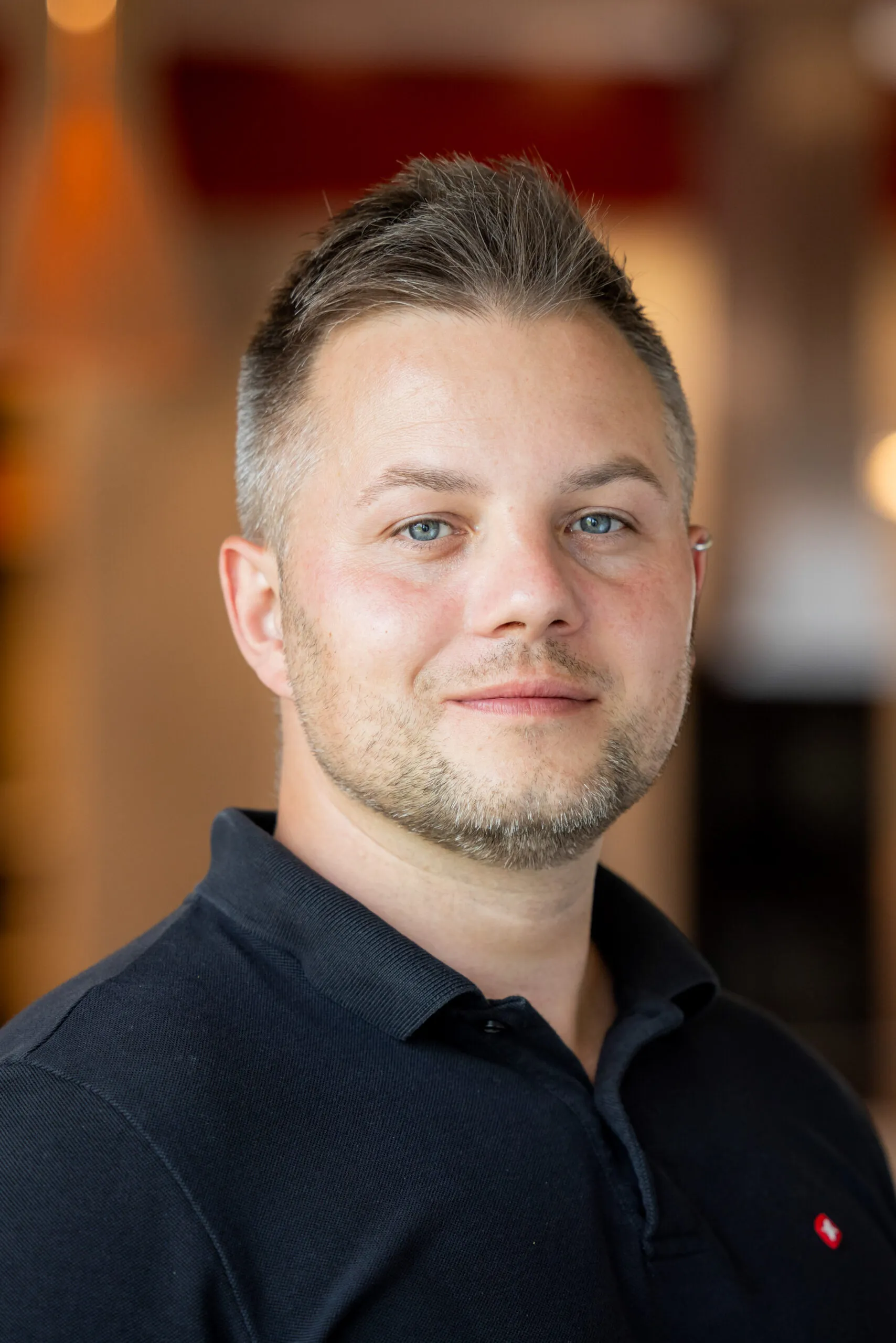 Ein sehr nah aufgenommenes Portraits von einem Mitarbeiter mit mittelbraunem, sehr kurzem Haar, Bart und einem Ohrring in seinem linken Ohr. Er lächelt ganz leicht und schaut in die Kamera, leicht gedrehte Körperhaltung. Er trägt ein dunkelblaues Polohemd. Der Hintergrund ist sehr unscharf in Orangen und Rottönen gehalten.
