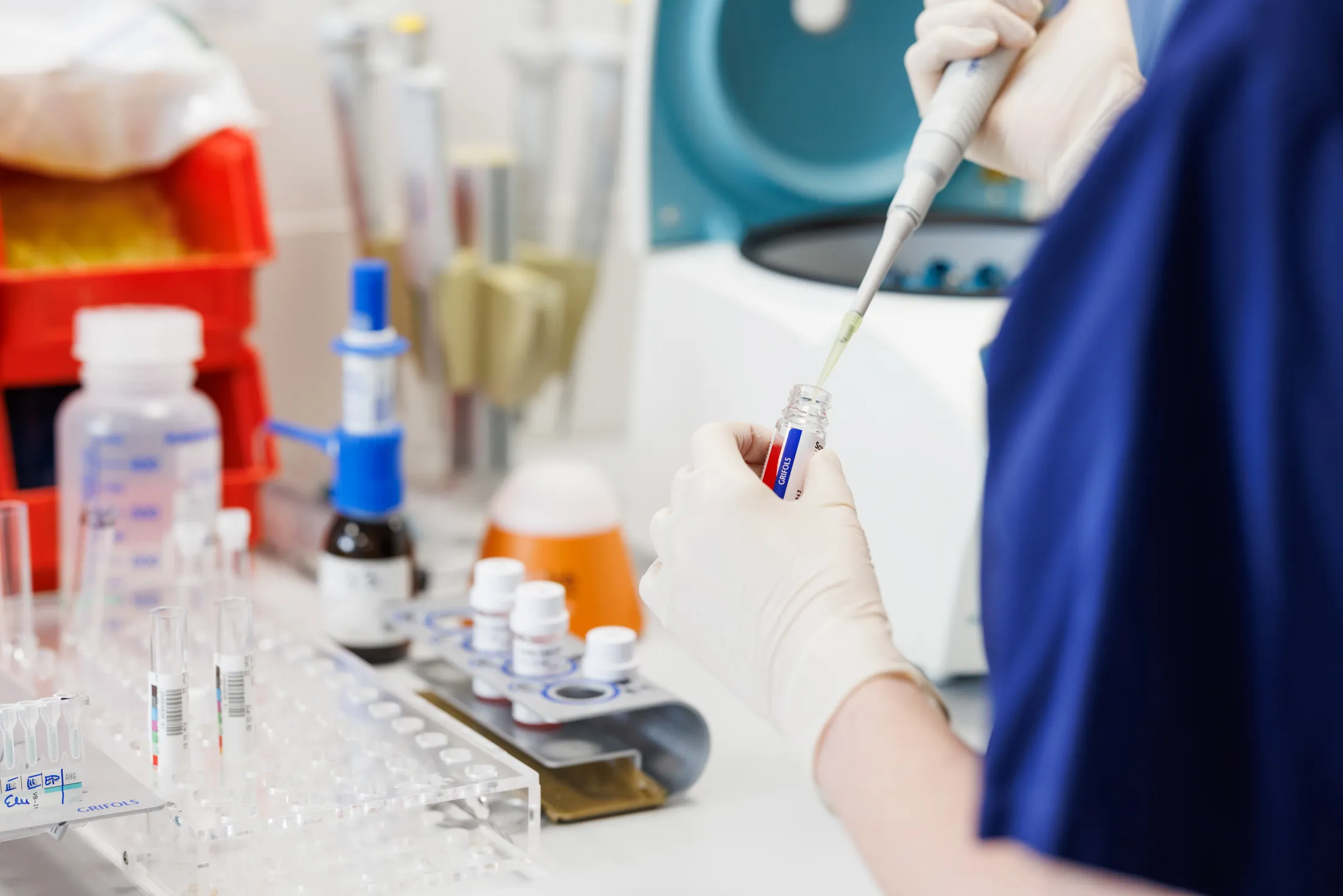 In einem Labor ist ein Cloesup auf zwei Hände mit weißen Handschuhen und Laborzubehör fotografiert. Eine Person ist rechts im Anschnitt nur mit einem dunkelblauen Kasack zu sehen, in der linken Hand hält sie ein Blutröhrchen und in der rechten Hand eine Pipette und tropft etwas in das Glas. Links sind auf einem Tisch weitere Blutproben, Reagenzgläser und Laborzubehör unscharf auf einem weißen Tisch zu sehen.
