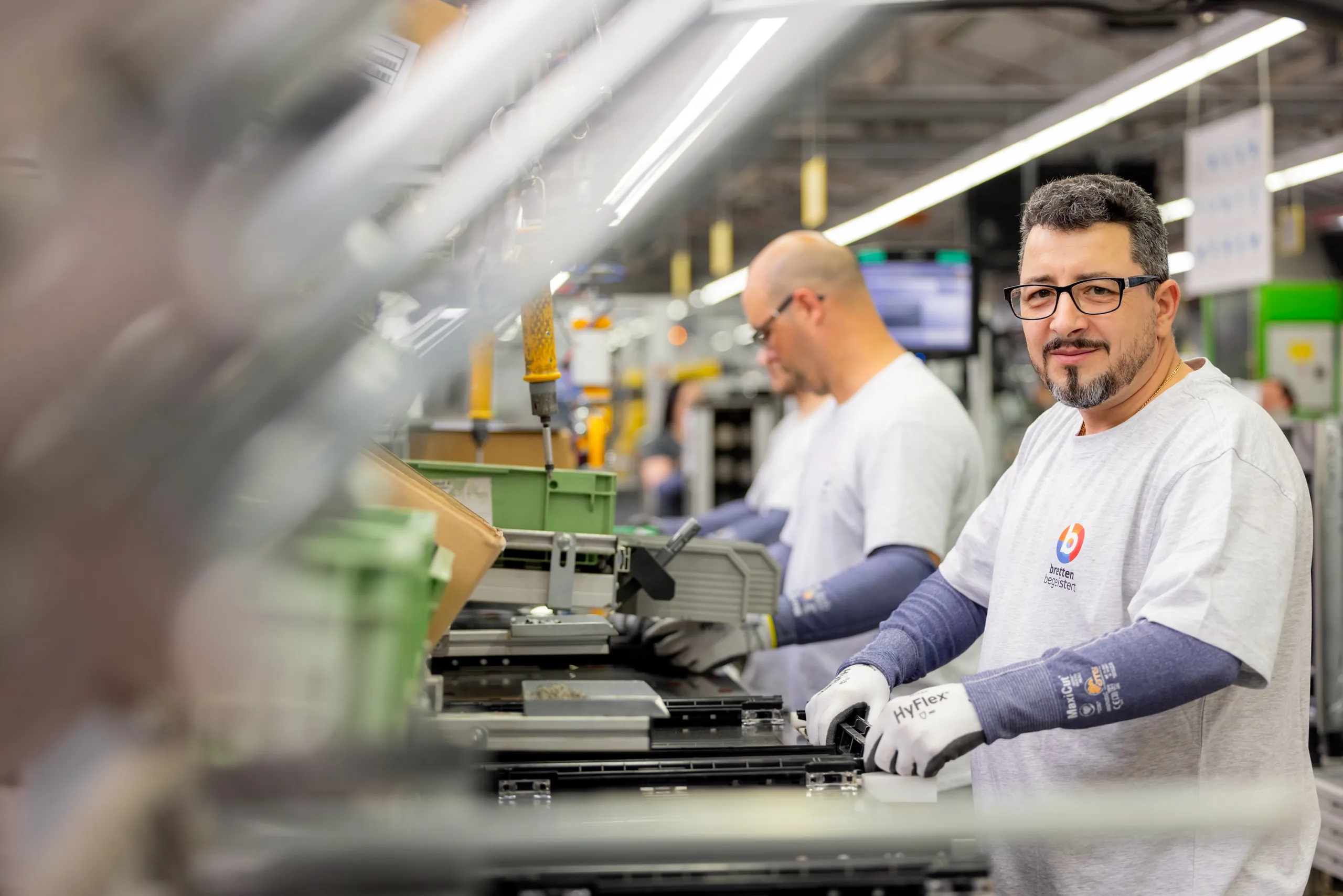 Drei Mitarbeiter stehen an einer Produktionslinie und arbeiten. Alle tragen ein hellgraues TShirt aus dem Werk, Sicherheitshandschuhe, die Hände und Arme bedecken. Der erste Mann hat graumelierte Haar, trägt einen Bart und eine dunkle Brille. Er schaut in die Kamera, die anderen sind unscharf und schauen auf ihre Arbeitsschritte