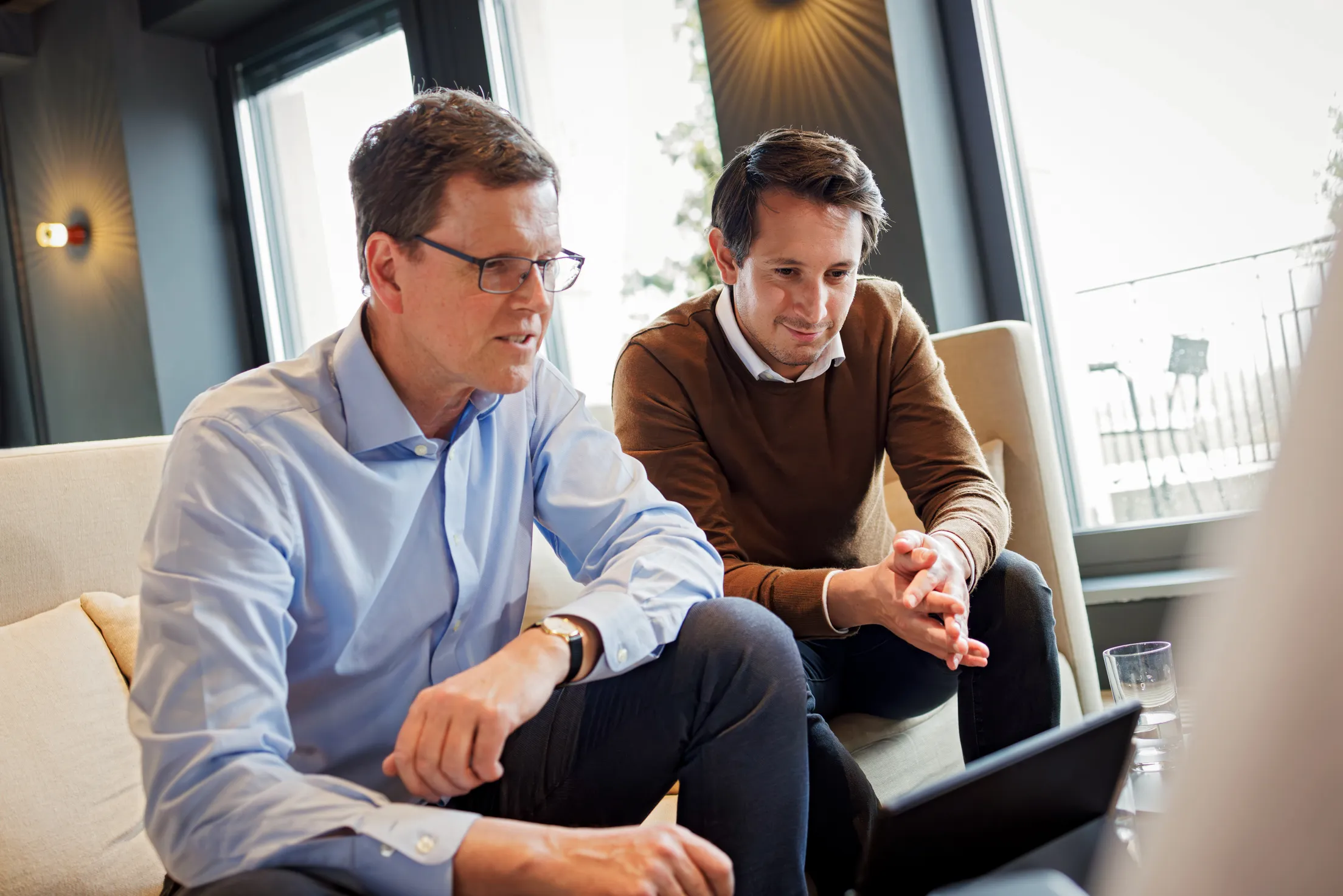 Zwei Männer sitzen auf einer hellen Couch und schauen in ein Tablet. Der Mann mittleren Alters links im Bild trägt eine graue Hose und ein hellblaues Hemd. Der andere jüngere Mann trägt ein weißes Hemd, darüber einen braunen Strickpullover und eine dunkle Hose. Im Hintergrund sind Glasfenster, graue Wände mit Wandleuchte und unscharf eine Dachterrasse erkennbar.