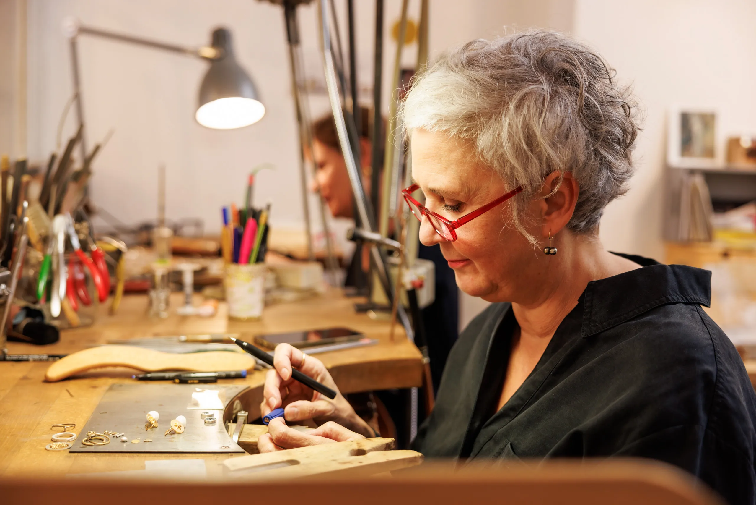 Die zweite Goldschmiedin von der Seite aufgenommen beim Bearbeiten eines Schmuckstückes in der Werkstatt. Sie hat graues, kurzes Haar, ein schwarzes Hemd und eine rote Brille. Im Hintergrund unscharf die Werksatt und die zweite Goldschmiedin am Arbeitsplatz zu sehen. Viel Werkzeug und eine Holzplatte mit auf dem Bild.