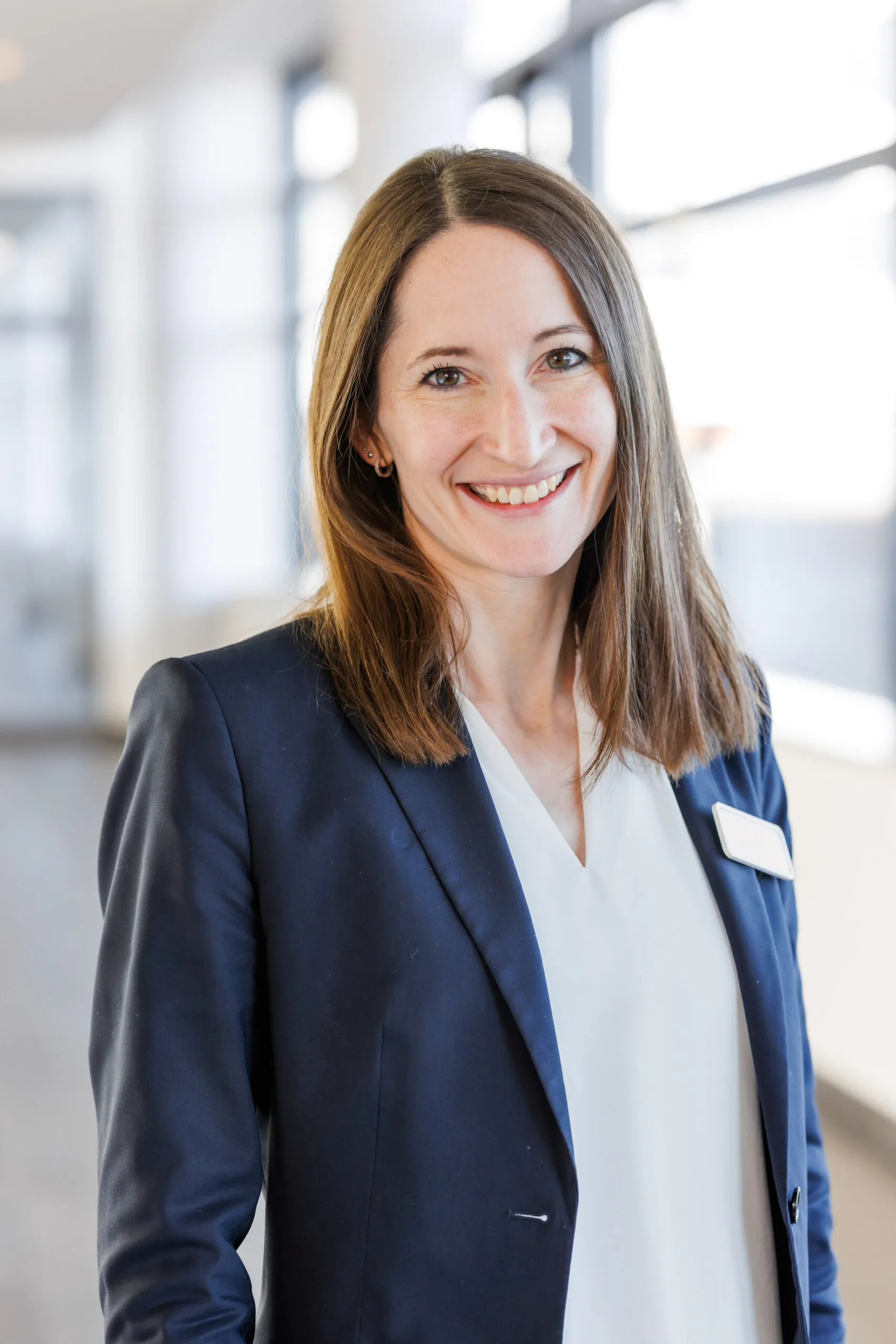 Hochformatiges Portrait eines jüngeren Klinikmitarbeiterin. Sie trägt einen dunkelblauen Blazer, ein weißes Shirt und hat braune schulterlange, glatte Haare. Sie lächelt in die Kamera und ist mit dem Oberkörper abgelichtet. Im Hintergrund ist unscharf ein heller Klinikgang mit Säulen und Fenster zu erkennen.