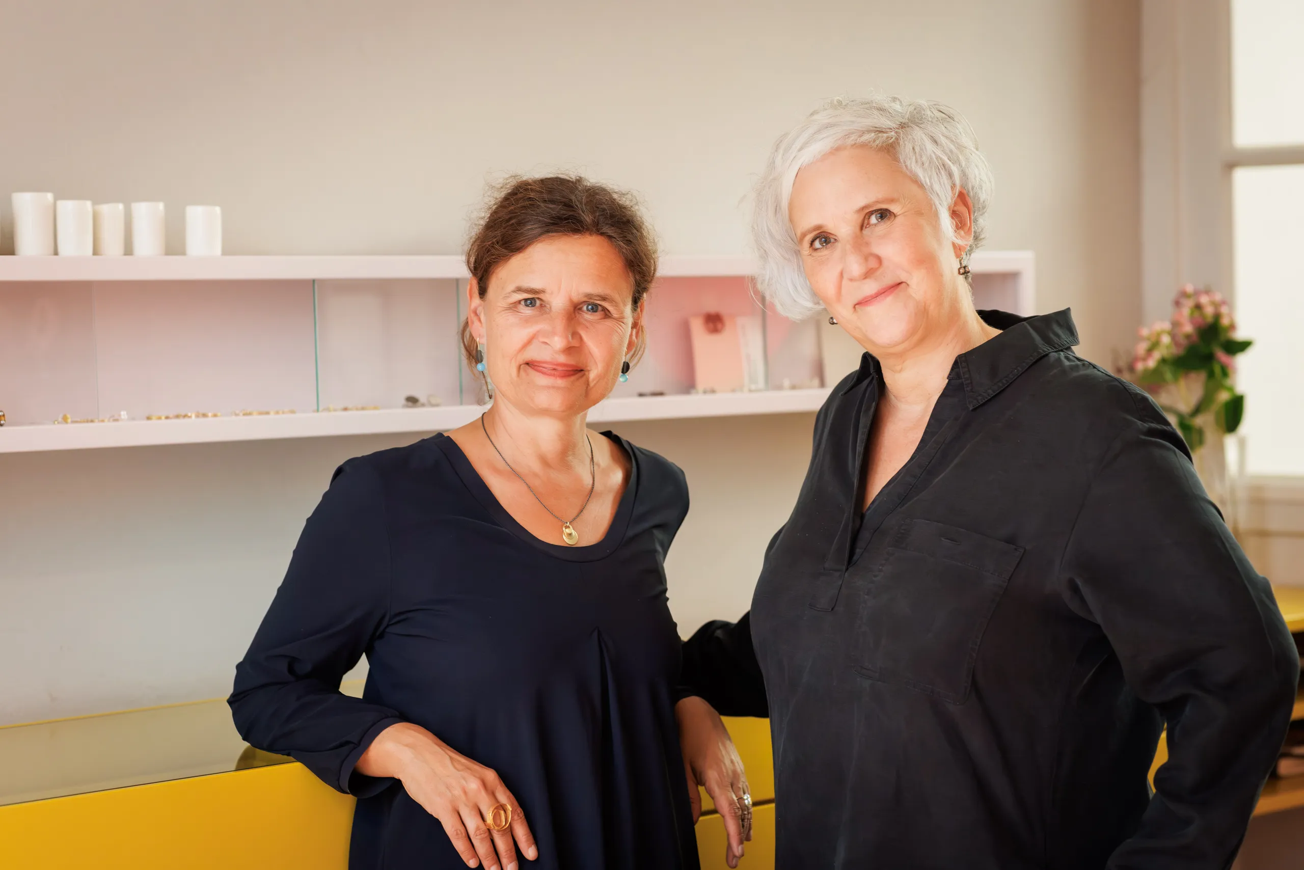 Zwei Goldschmiedinnen stehen nebeneinander vor einer gelben Vitrine und einer weißen Wand mit zwei Regalen. Rechts ein Blumenstrauss zu sehen. Beide tragen Schmuck, dunkle Kleider und schauen in die Kamera. Die Frau links hat ihr braunes Haar zurückgebunden und die Frau rechts im Bild hat graues, kurzes Haar.