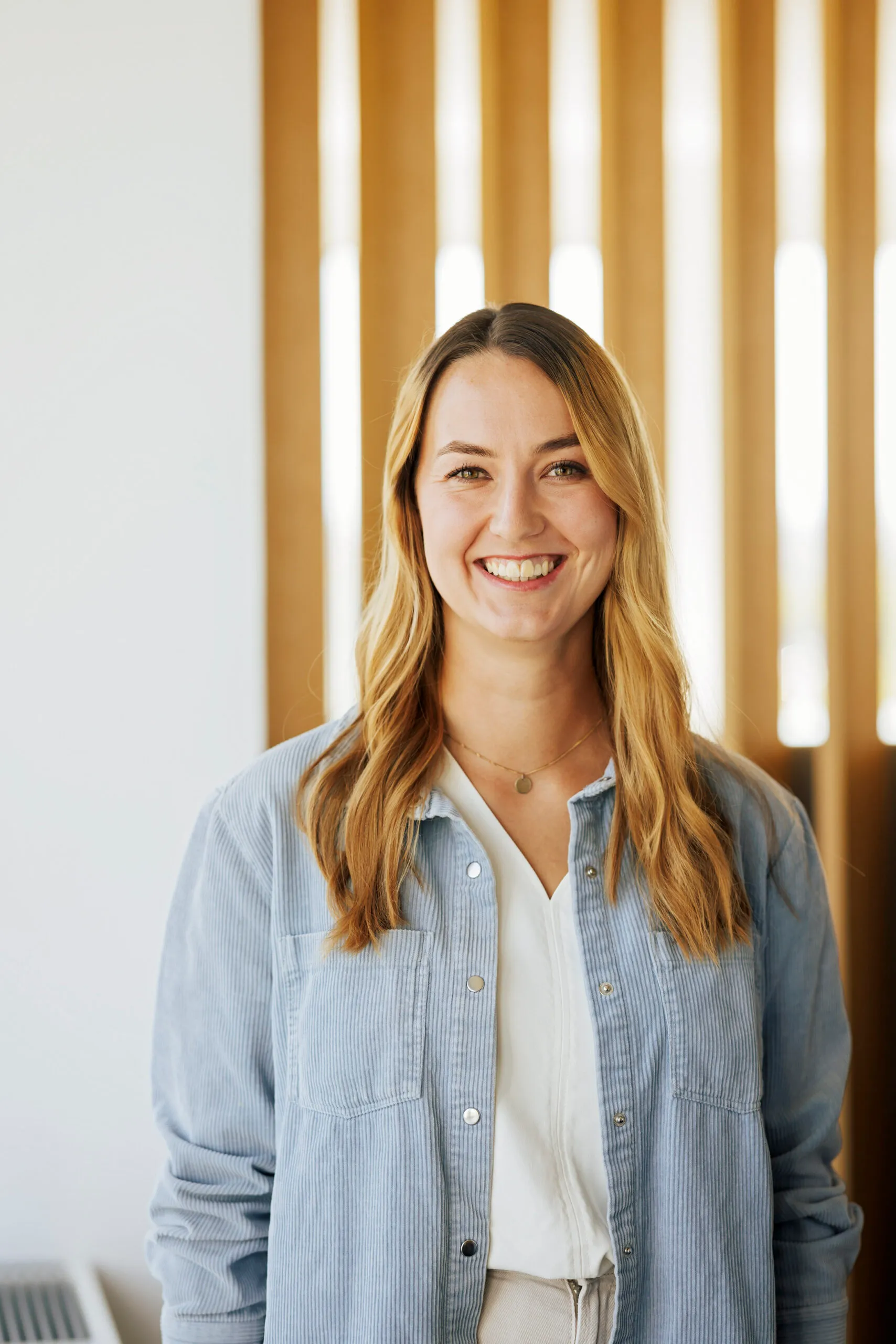 Eine junge Frau mit mittelblondem langem Haar steht mit einem hellen Jeanshemd vor einer gestreiften Holzwand und schaut in die Kamera