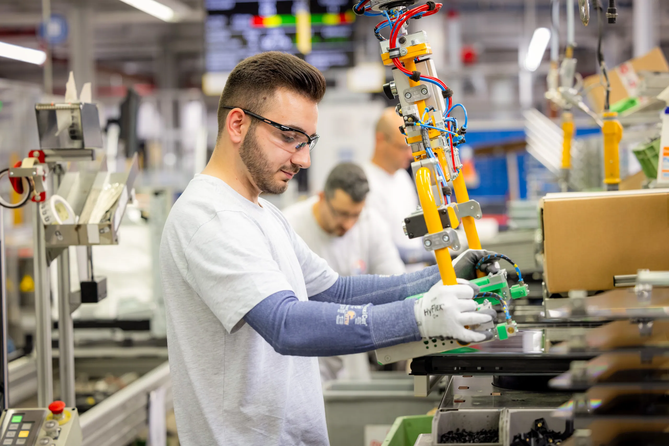 Ein junger Mann mit braunem Haar, Bart trägt ein hellgraues T-Shirt, Sicherheitsbrille und blaue Sicherheitshandschuhe bis hoch zu den Oberarmen. Er steht an einem Gerät und hat einen Greifarm der Maschine in der Hand. Im Hintergrund unscharf zwei weitere Männer beim Arbeiten an der Produktionslinie mit vielen Geräten.