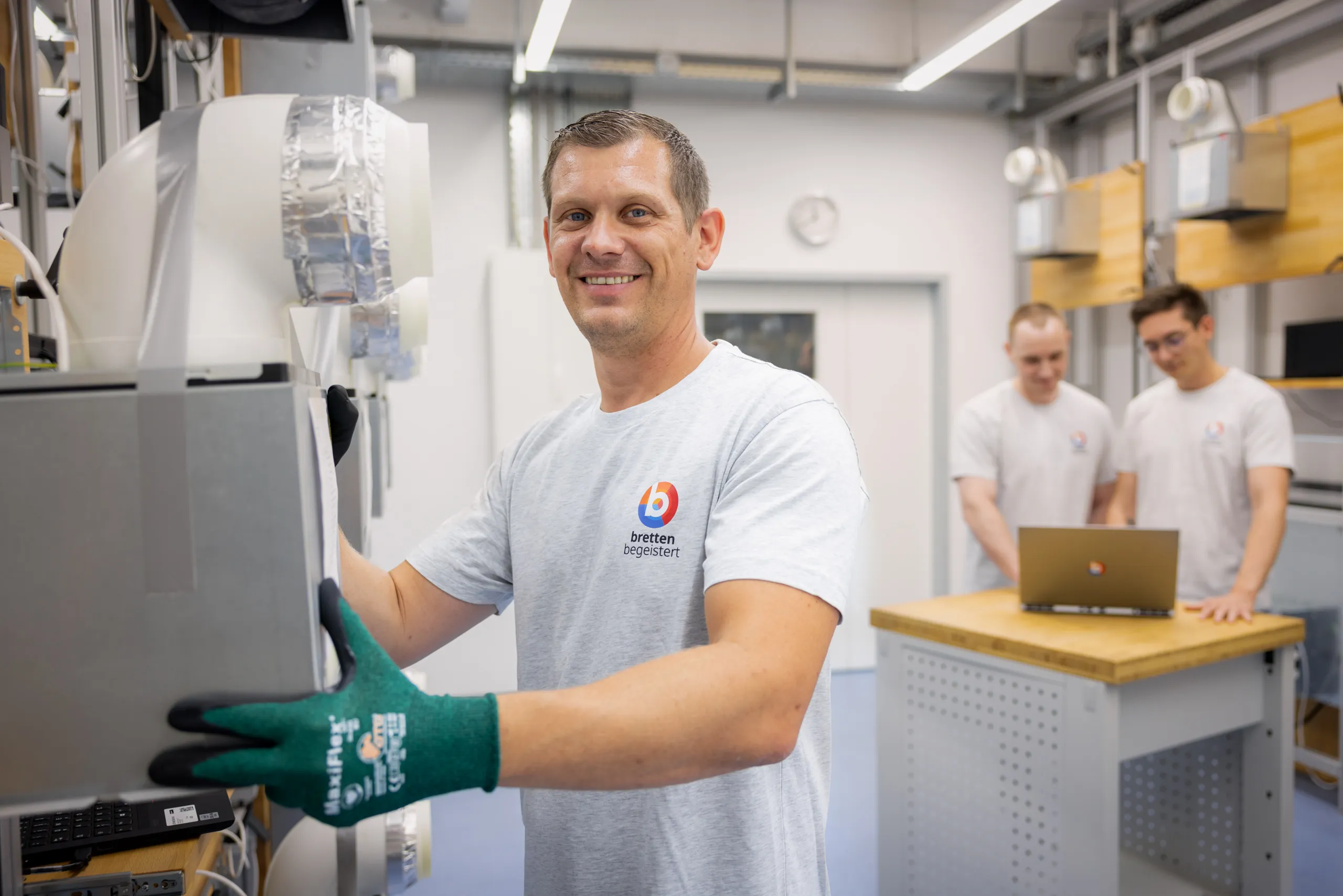 In einem sehr hellen Raum der Entwicklung sind drei Männer auf dem Foto. Links vorne im Bild ist scharf abgebildet ein junger Mann mit einem grauen BSH T-Shirt und grünen Sicherheitshandschuhen. Er hält eine eingepackte Dunstabzugshaube mit beiden Händen fest. Rechts hinten im Bild stehen in der Unschärfe zwei junge Männer mit hellgrauem Shirts und sie schauen in ein Laptop, welcher auf einem kleinen Podest mit einer Holzplatte steht. Im Hintergrund hängen noch andere Abzugshauben an der Wand.