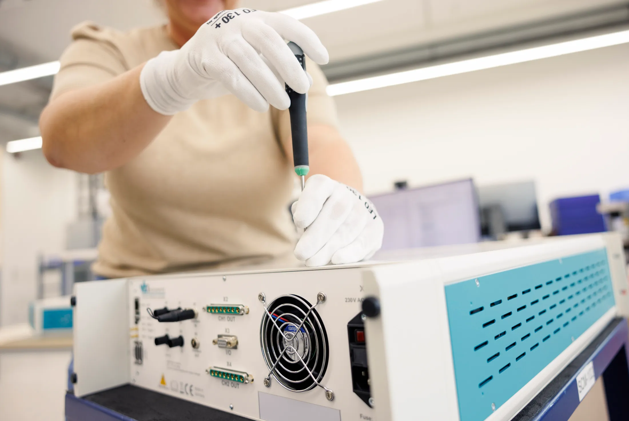 Zwei Hände mit Handschuhe schrauben ein Halbleiter Testsystem mit einem Schraubenzieher auf. Es ist unscharf der Oberkörper eine Frau mit beigem Shirt zu sehen.