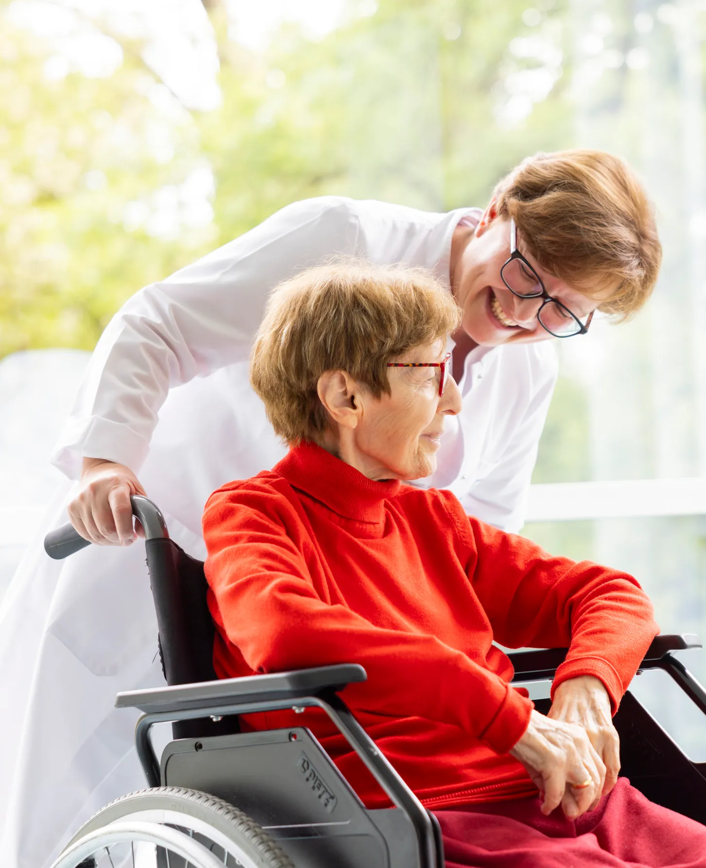 Eine Ärztin schiebt einen Rollstuhl und beugt sich sehr fürsorglich zu der Patientin im Rollstuhl. Diese trägt einen knalligen roten Rollkragenpullover und eine rote Brille.